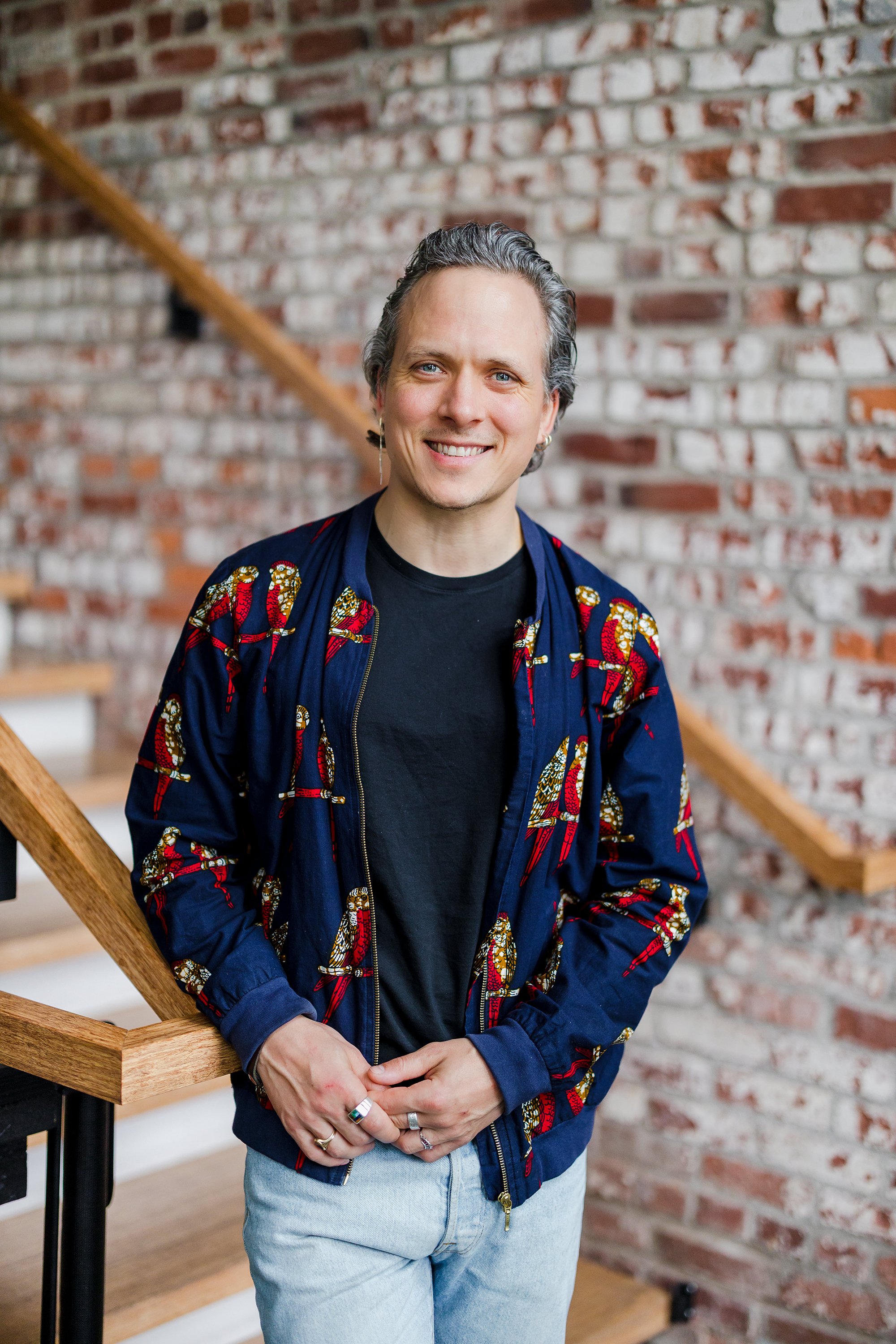 A person with short wavy gray hair, wearing a blue jacket with colorful snake patterns, a black shirt, and light blue jeans, standing in front of a brick wall with a wooden staircase and handrail in the background, smiling at the camera.