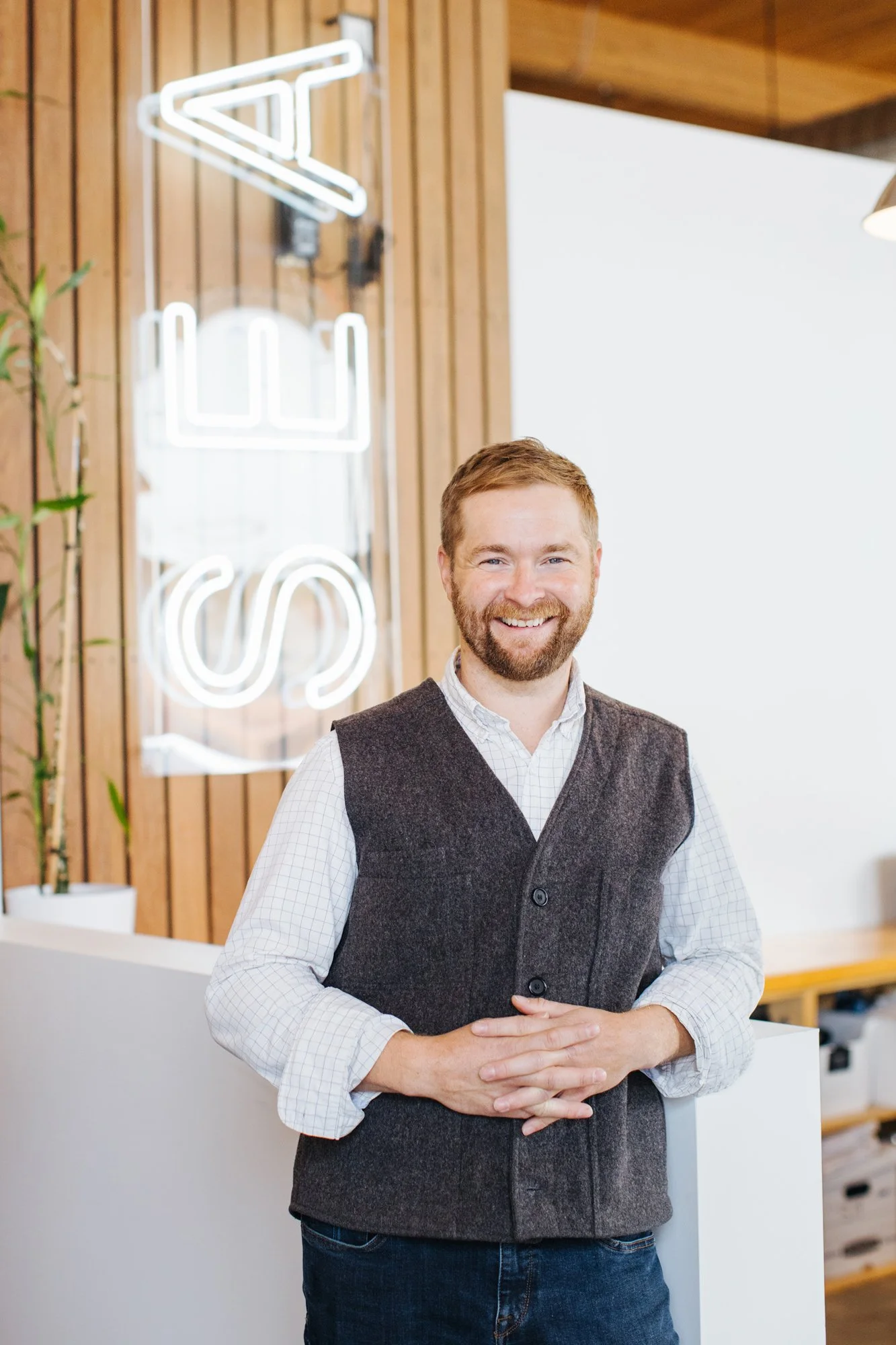 Smiling man in a checkered shirt and gray vest standing indoors near a wooden wall with a neon sign that reads "SIR" and a potted plant.