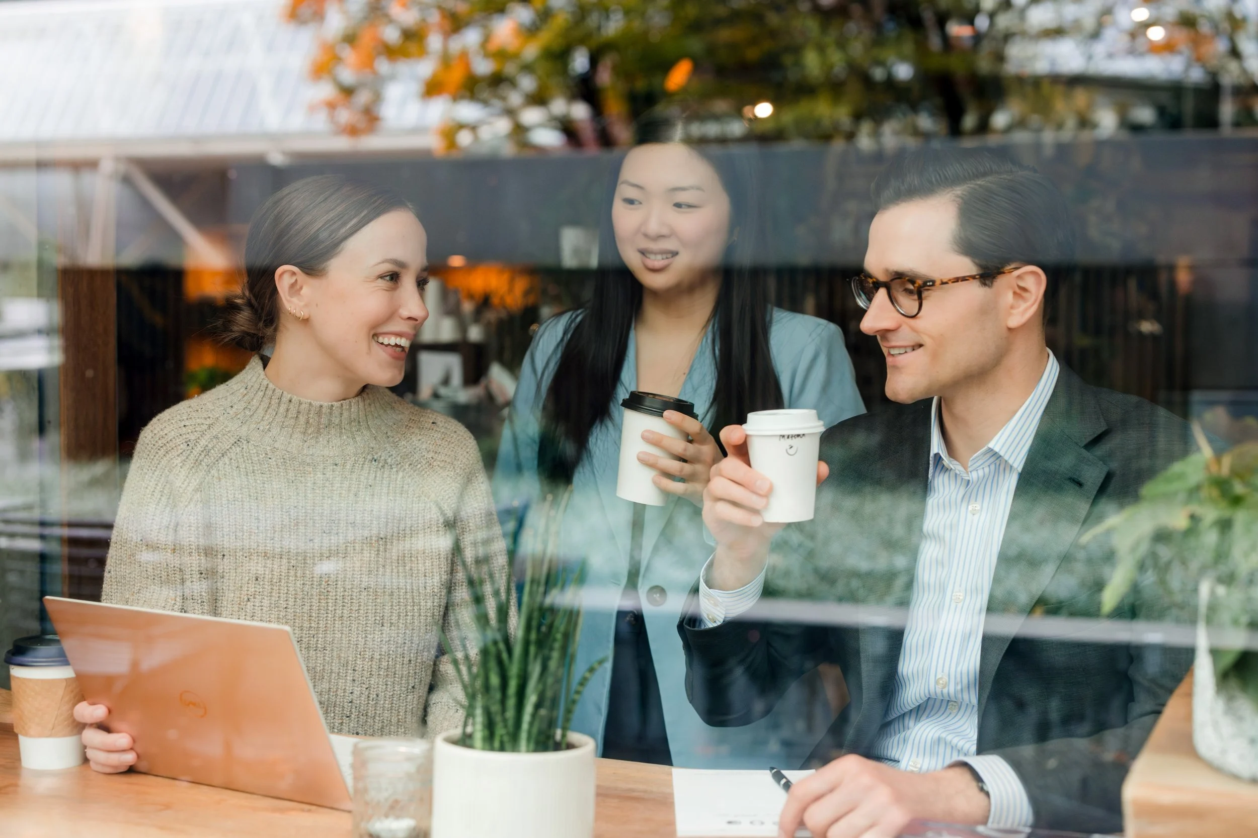 Three people, two women and one man, sitting at a table in a cafe, engaged in conversation. They are holding coffee cups and smiling. The photo is taken through a window with reflections of the outdoor scene.