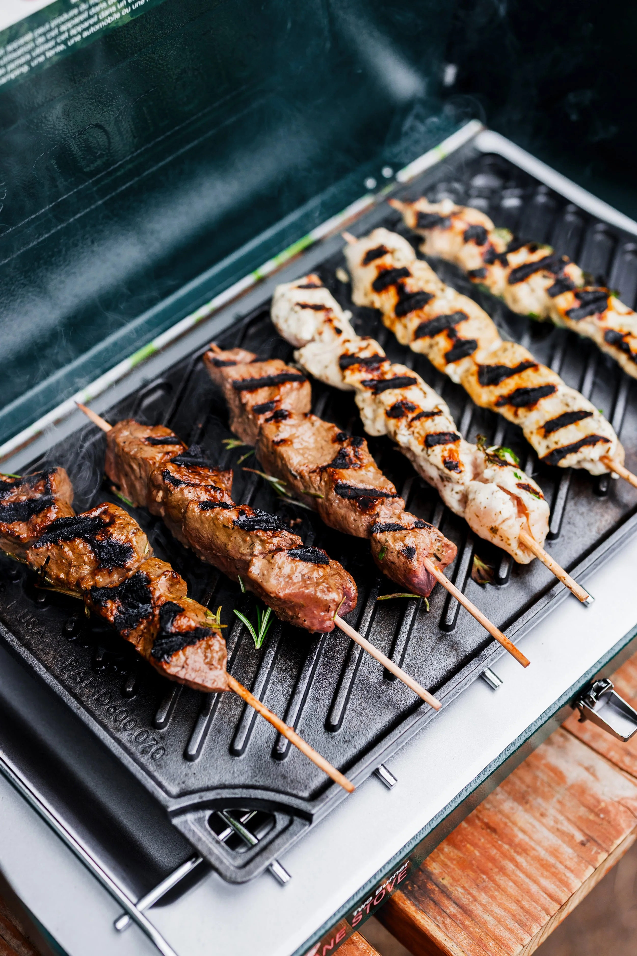 Assorted grilled kebabs on a barbecue grill with grill marks, including beef, chicken, and veggie skewers, with a pink surface.