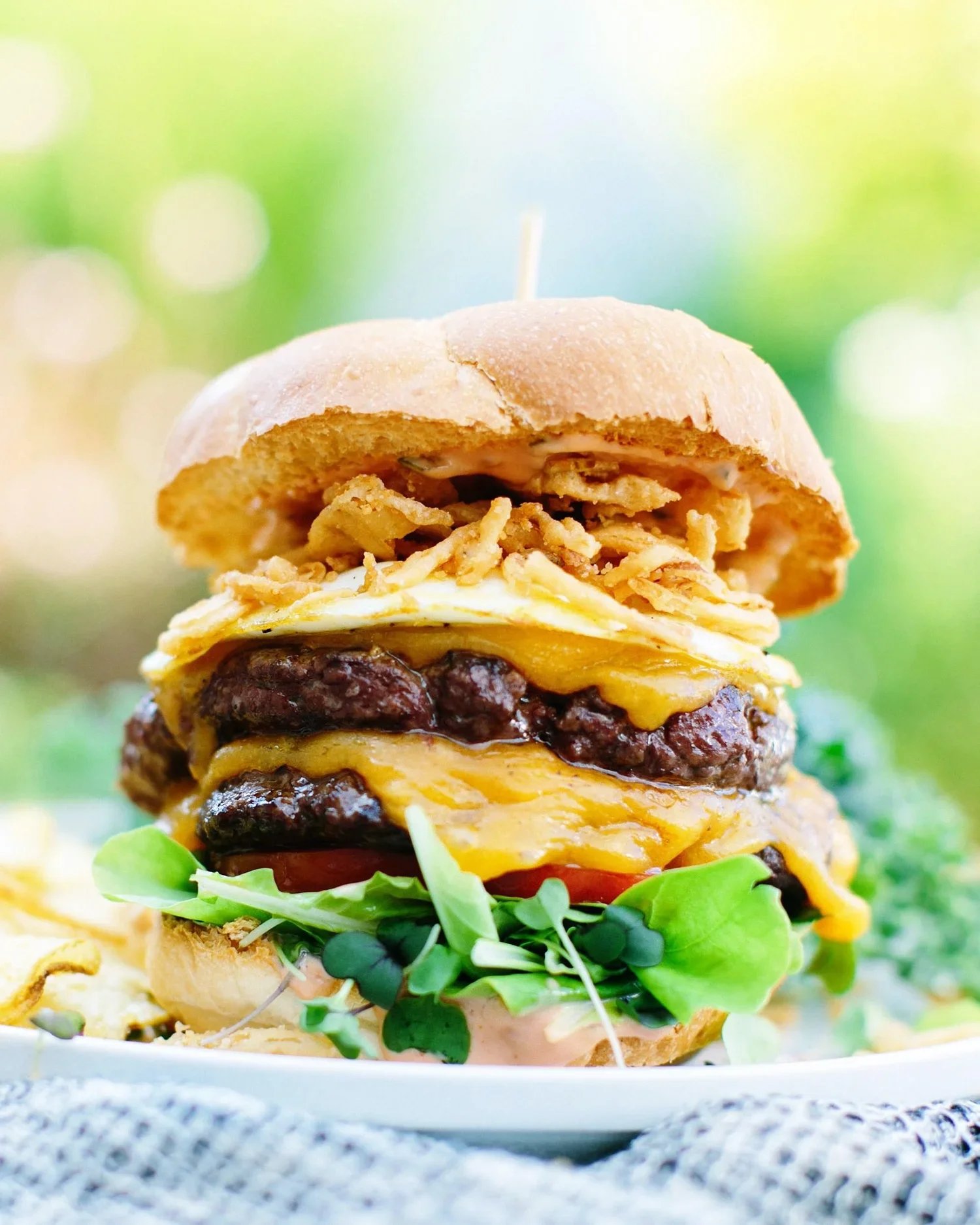 Close-up of a multi-layered cheeseburger with lettuce, tomato, cheese, beef patties, fried onions, a fried egg, and a sandwich bun, with a green outdoor background.