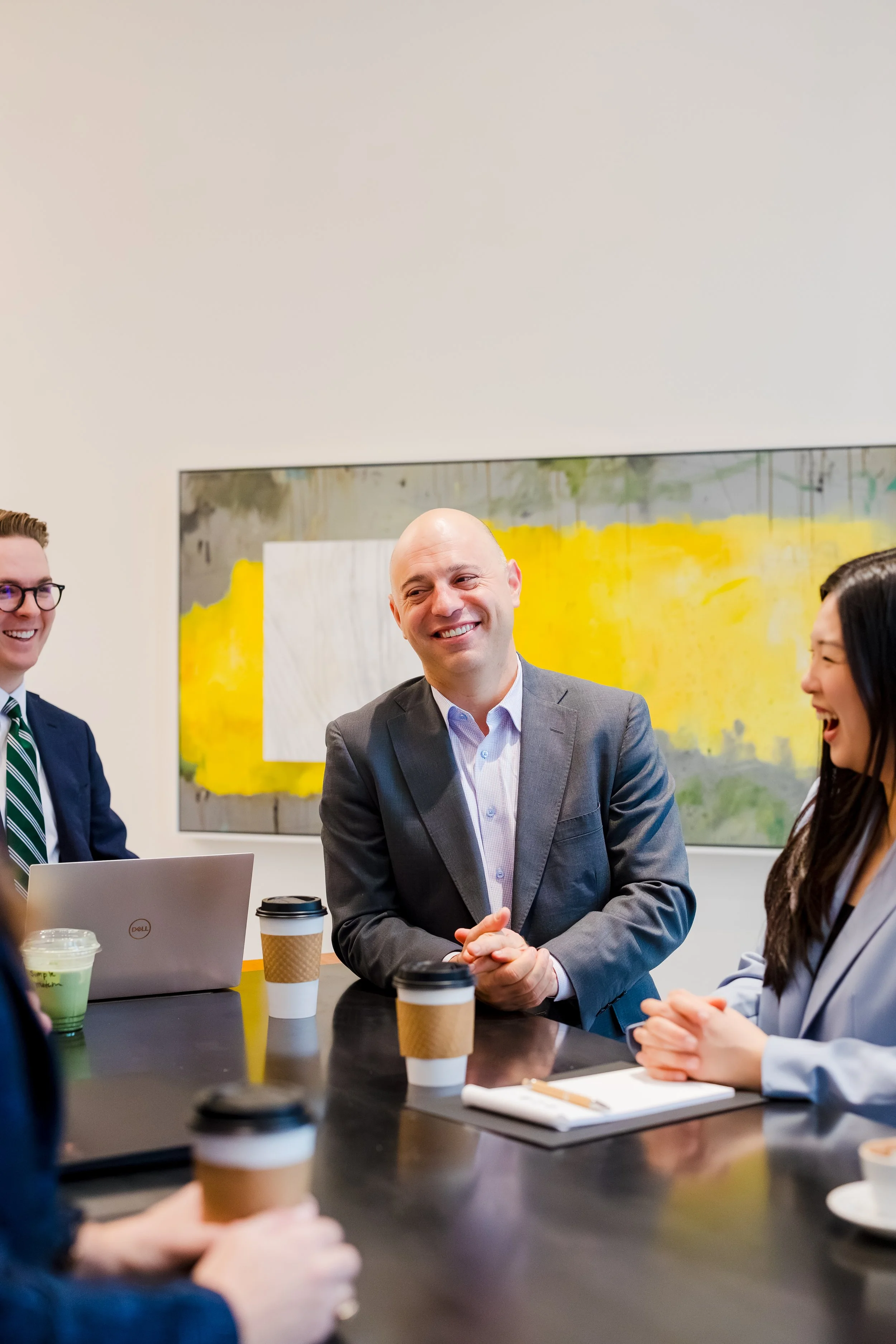 Group of businesspeople having a meeting in a conference room with coffee cups, a laptop, and a notepad, smiling and engaging in conversation.