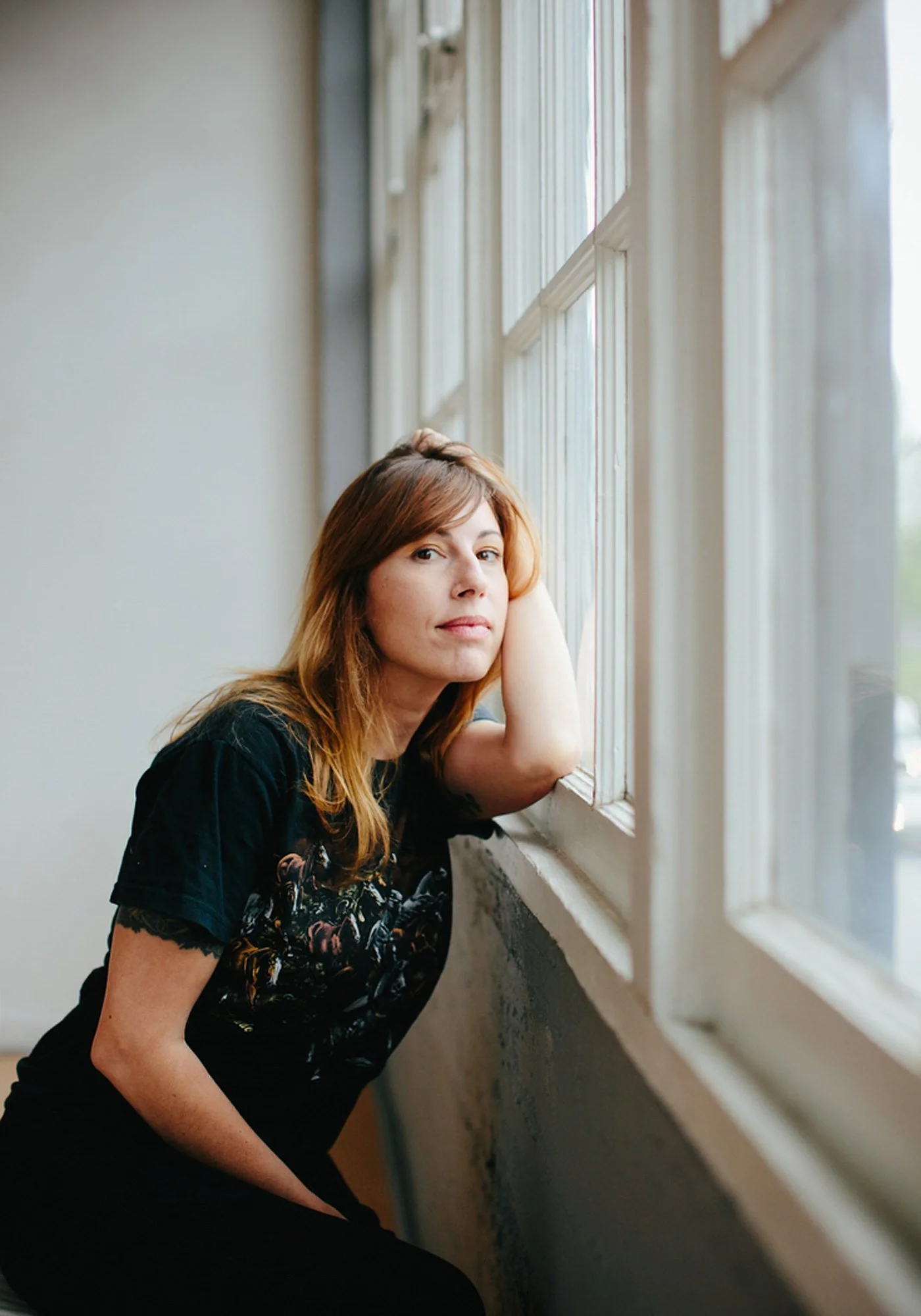 A woman with shoulder-length brown hair, wearing a black T-shirt, is leaning on her hand and looking out a multi-pane window.