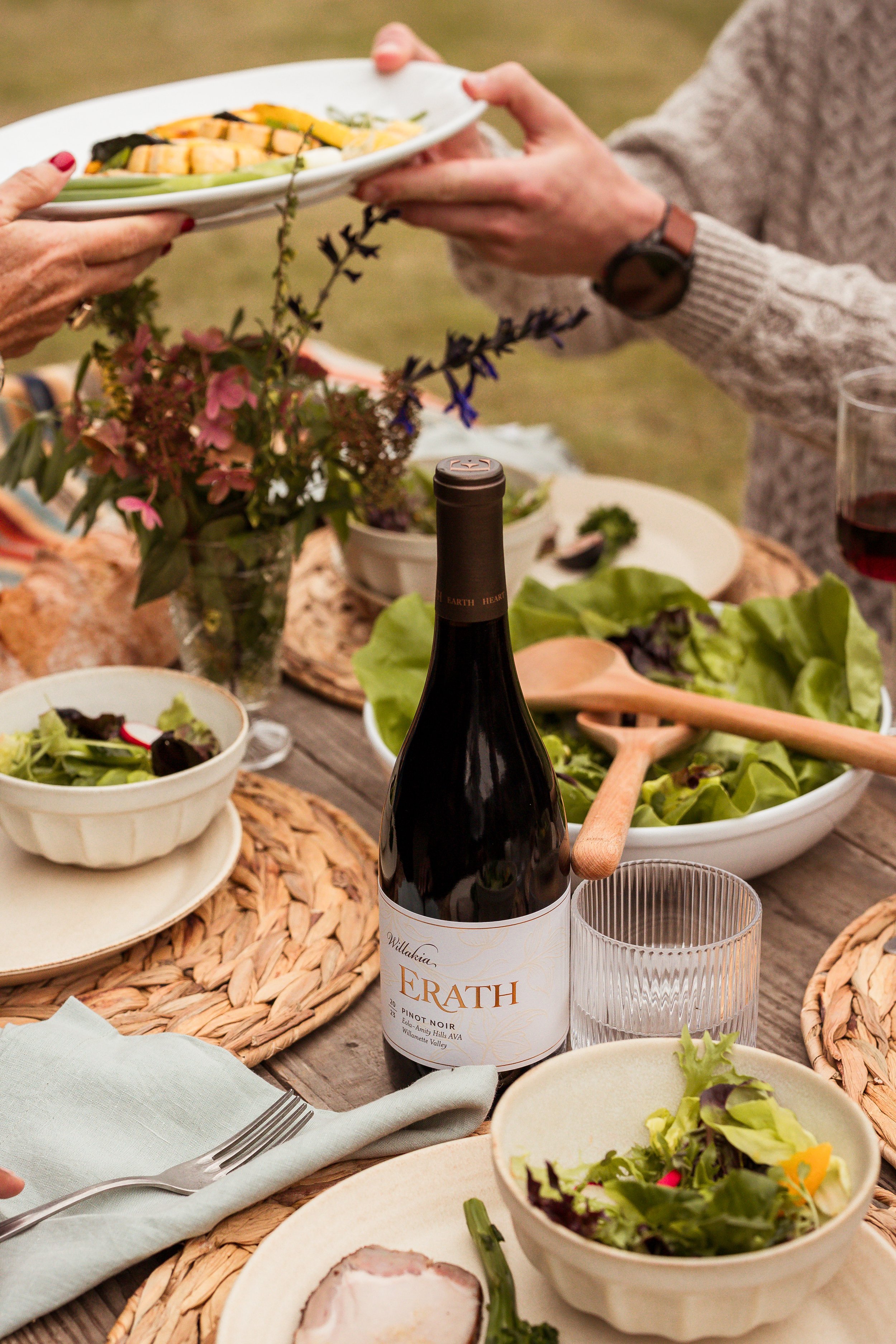 A wine bottle labeled 'Erath', a glass of red wine, bowls of salad, bread, and a person handing a plate of food at an outdoor dining table with flowers.