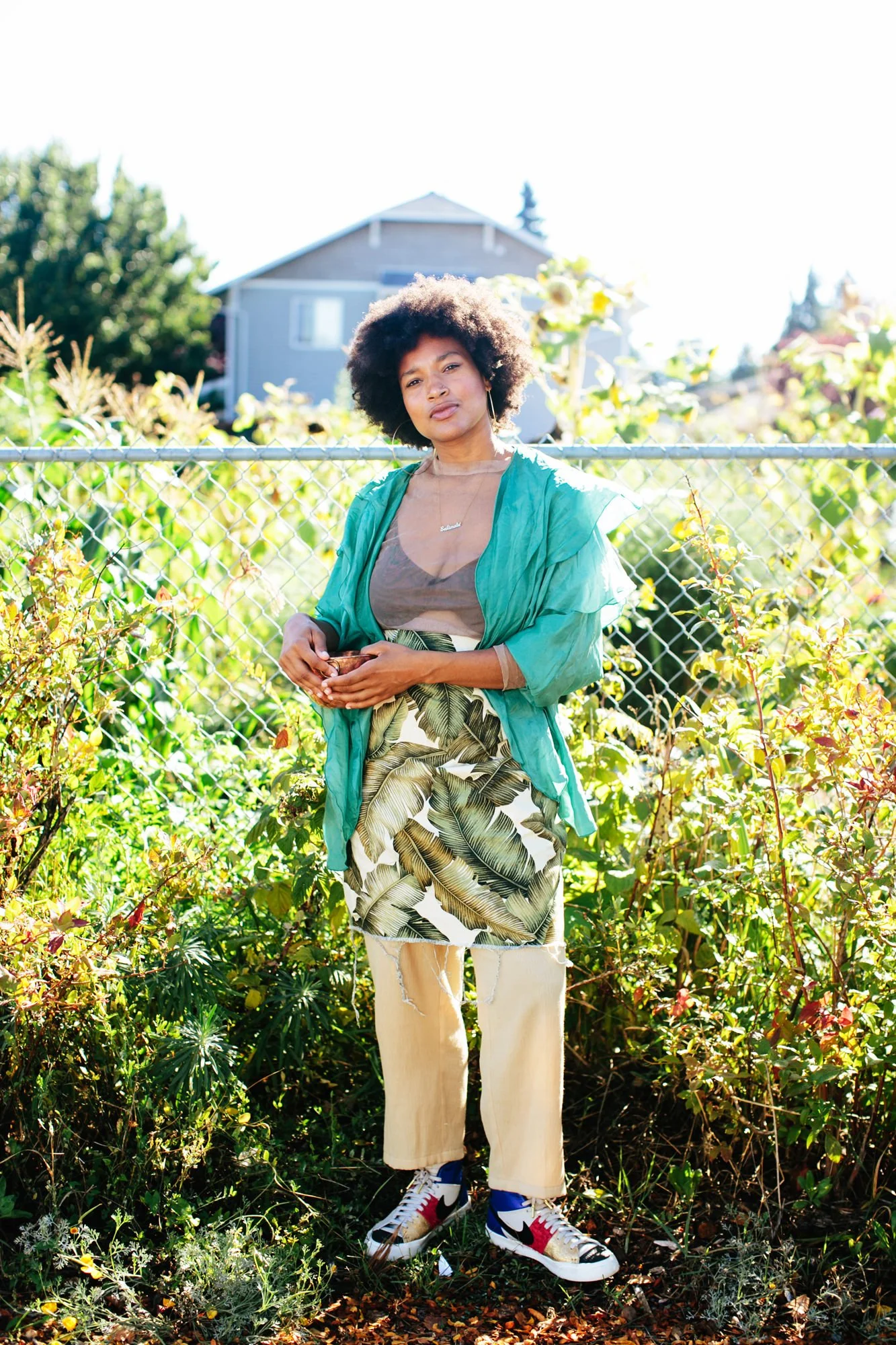 A woman with curly hair standing outdoors in front of a chain-link fence, surrounded by lush greenery and plants, with a house in the background, wearing colorful clothes and sneakers.