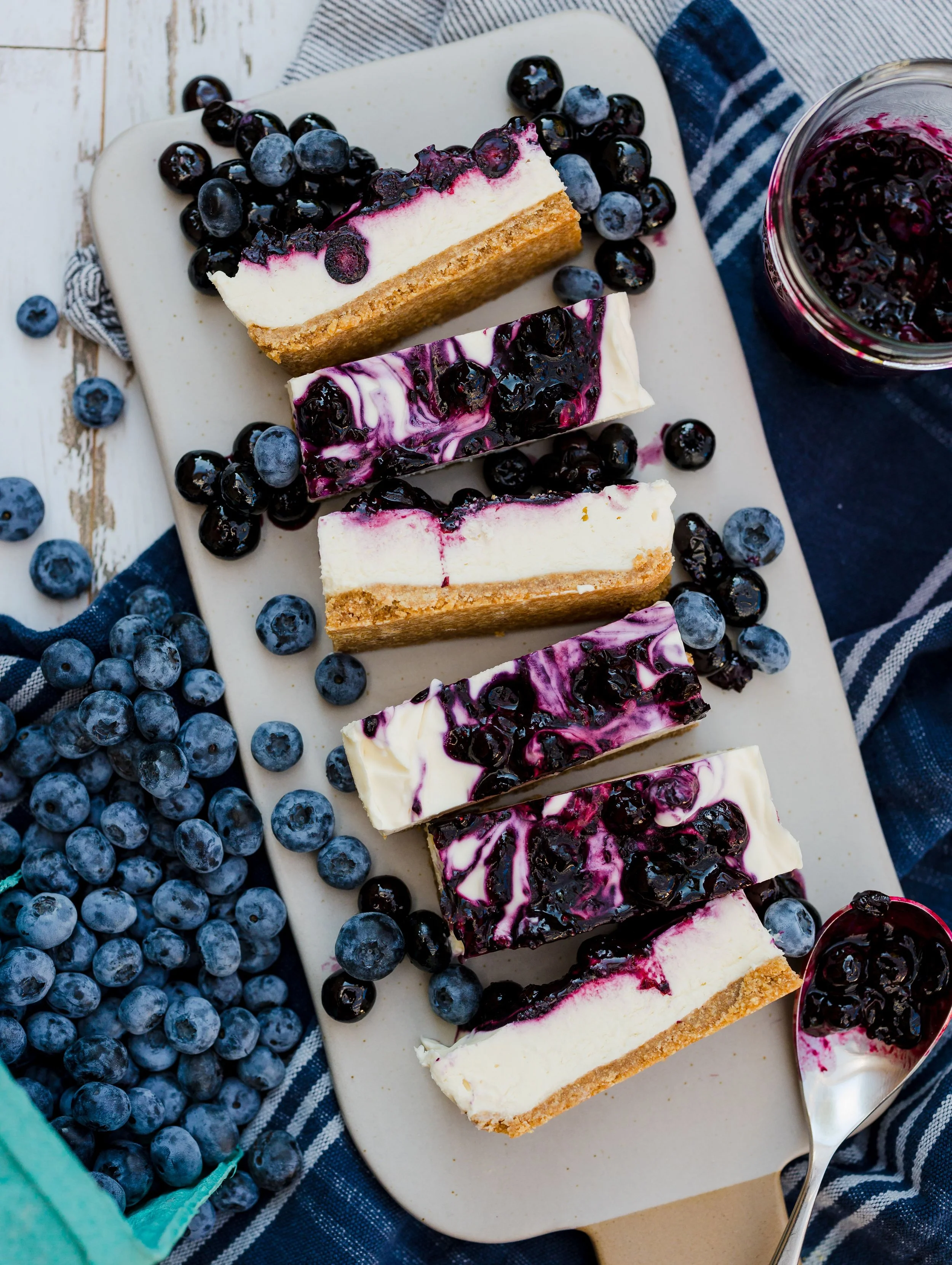 Slices of blueberry cheesecake on a white cutting board, surrounded by fresh blueberries and a jar of blueberry compote.