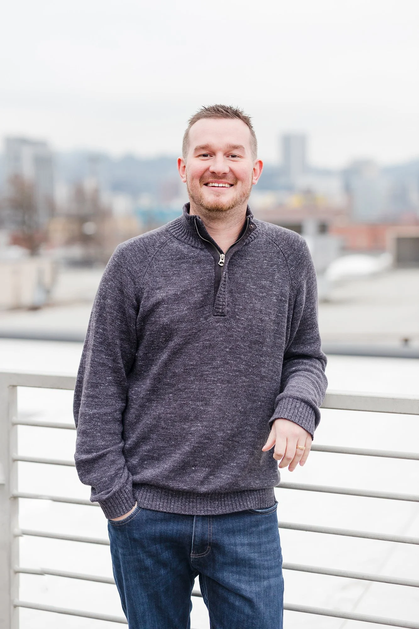 A smiling man with short hair and a beard wearing a dark gray sweater standing outdoors near a white railing with a city skyline in the background.