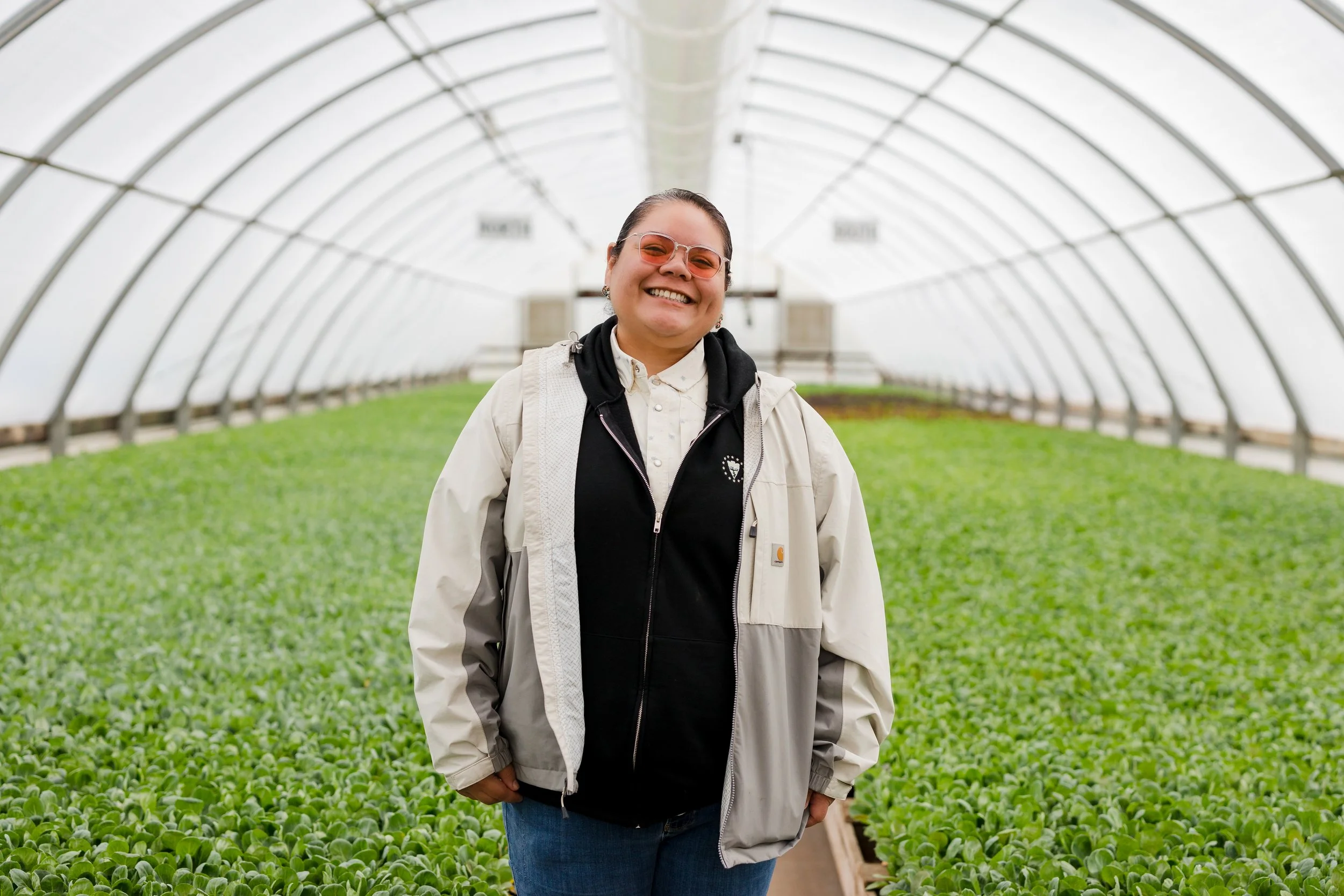 A smiling woman standing inside a greenhouse filled with leafy green plants.