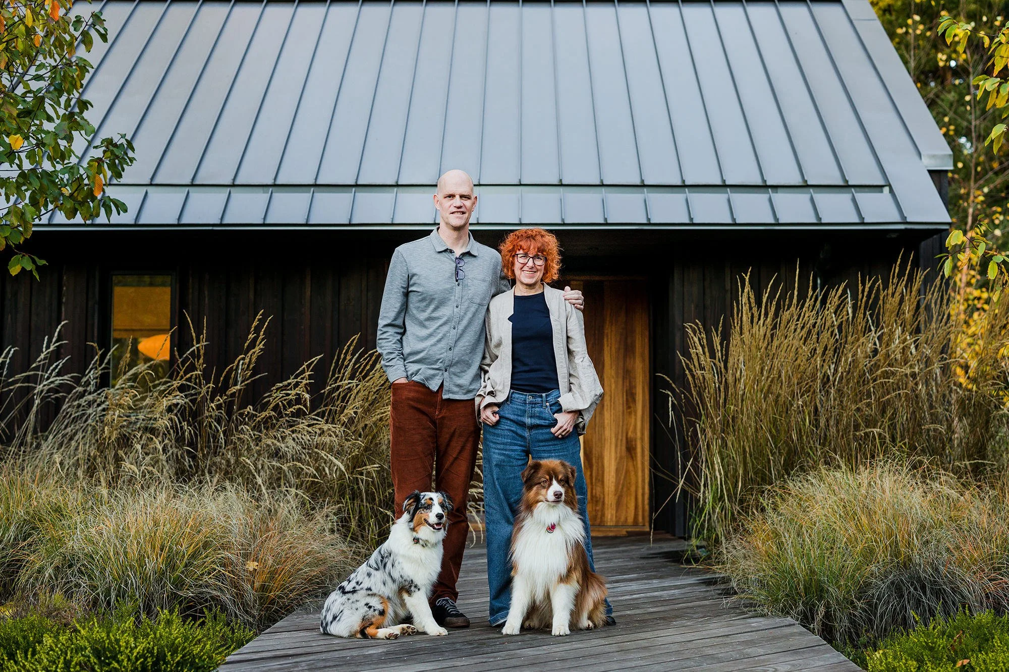 A couple with two dogs standing on a wooden deck outside a black house with a metal roof, surrounded by tall grasses.