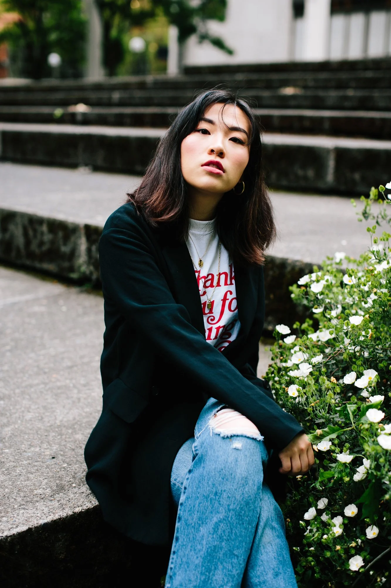 A young woman sitting on a sidewalk curb next to white flowering plants, with stone steps in the background.