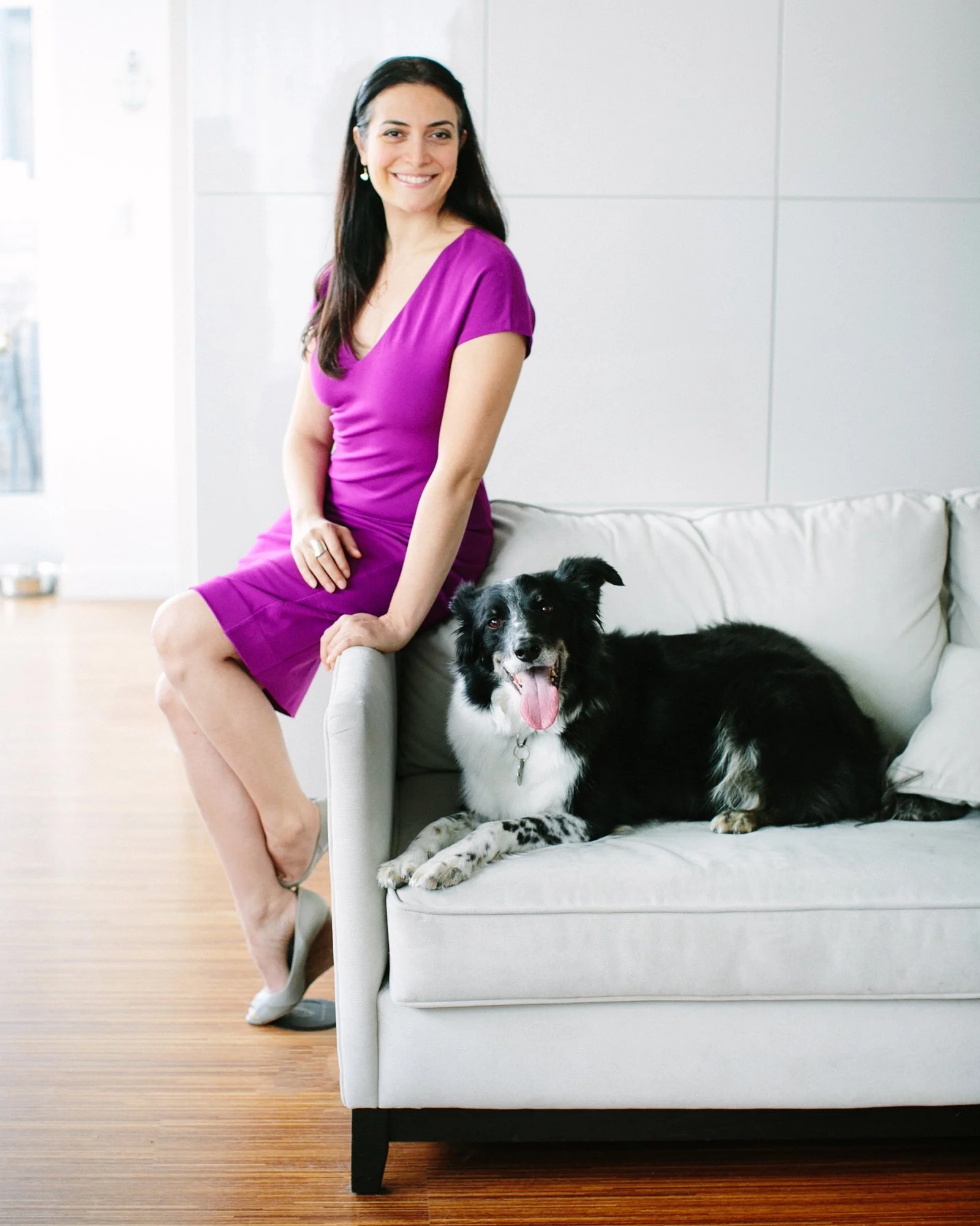 A woman in a purple dress sitting on a white sofa with a black and white dog lying beside her.