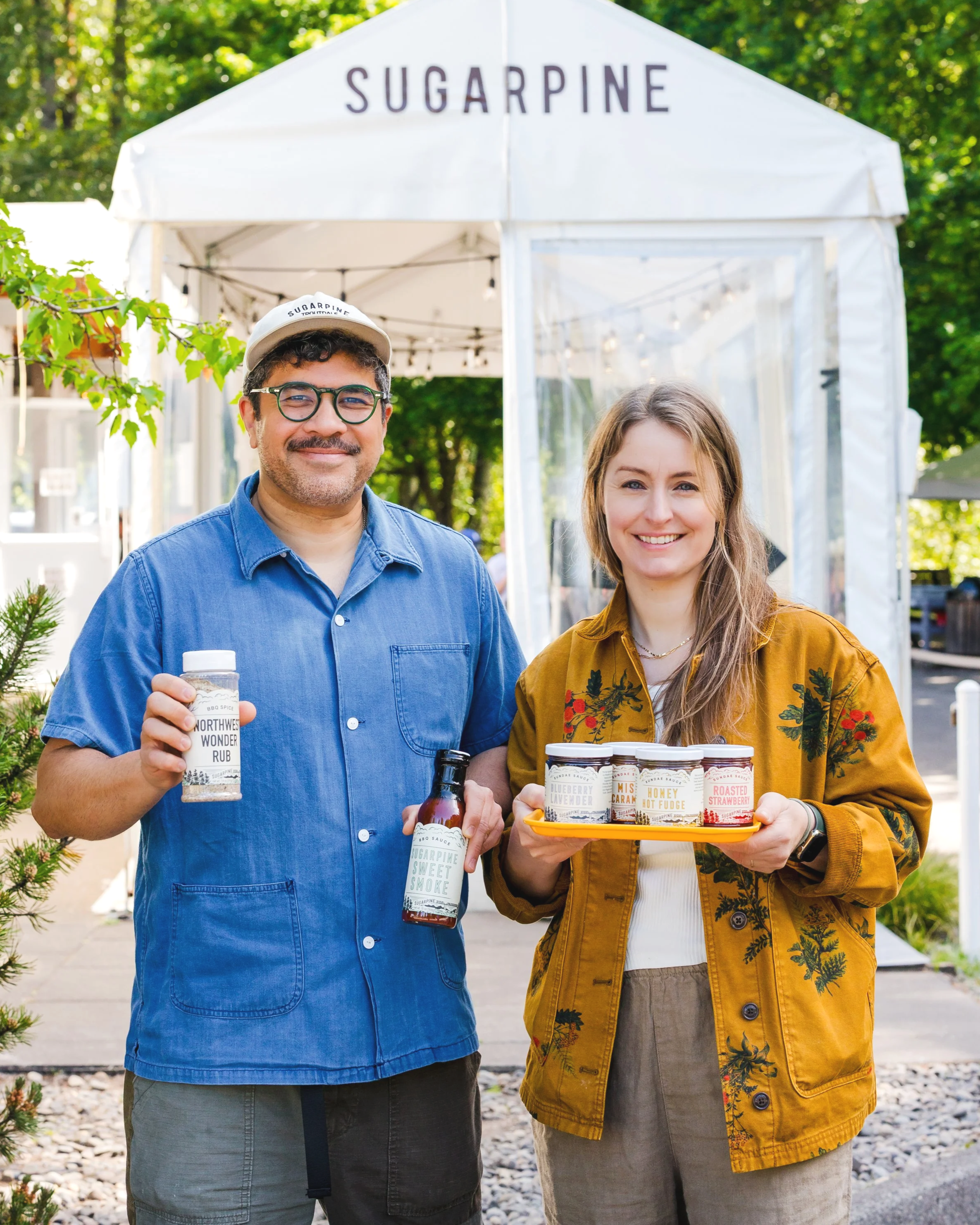 Two smiling people holding jars of jam and a bottle of sauce standing outside in front of a white tent with the word 'Sugarpine' on it, surrounded by trees.