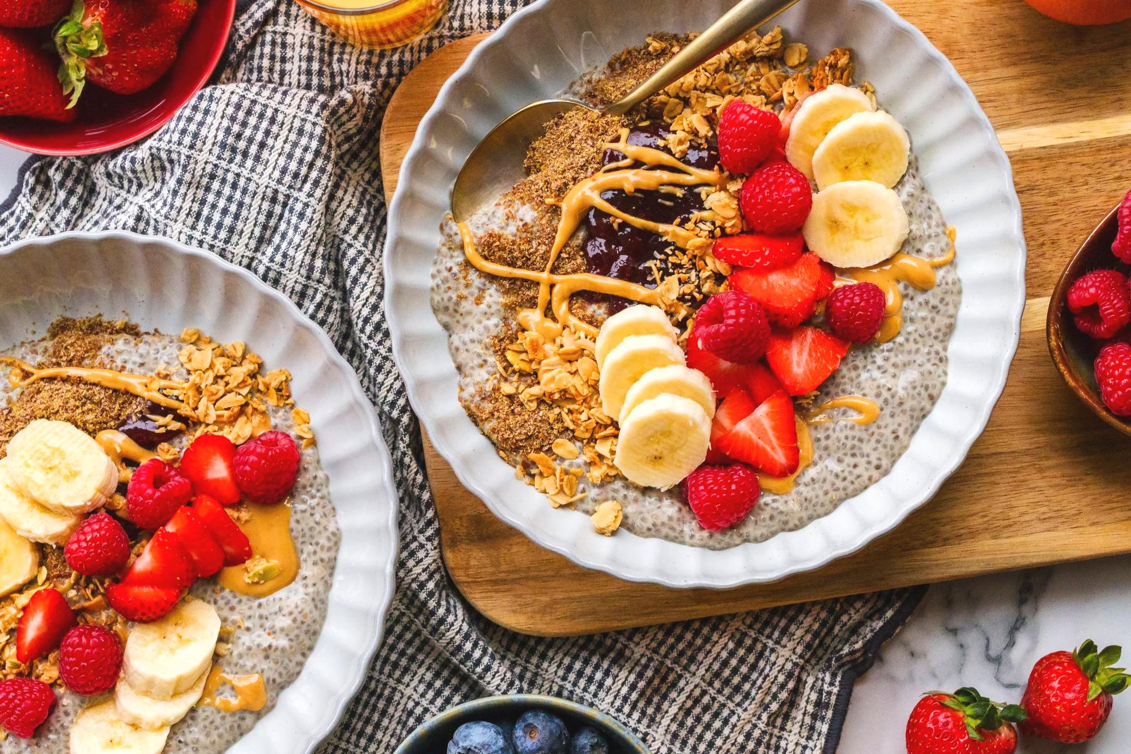 Overhead view of chia seed pudding topped with banana slices, strawberries, raspberries, almond flakes, and drizzled with peanut butter and chocolate sauce on a white fluted plate, with additional bowls of berries nearby.