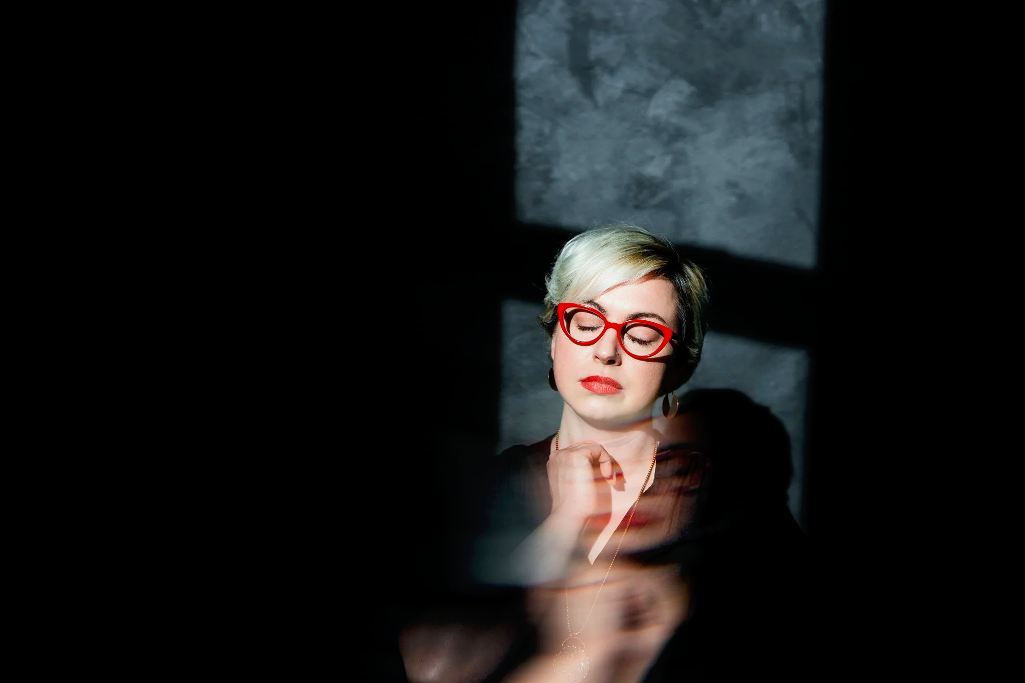 A woman with short blonde and brown hair wearing red glasses, closed eyes, and a black top, is illuminated by a window's shadow on a gray textured wall behind her.