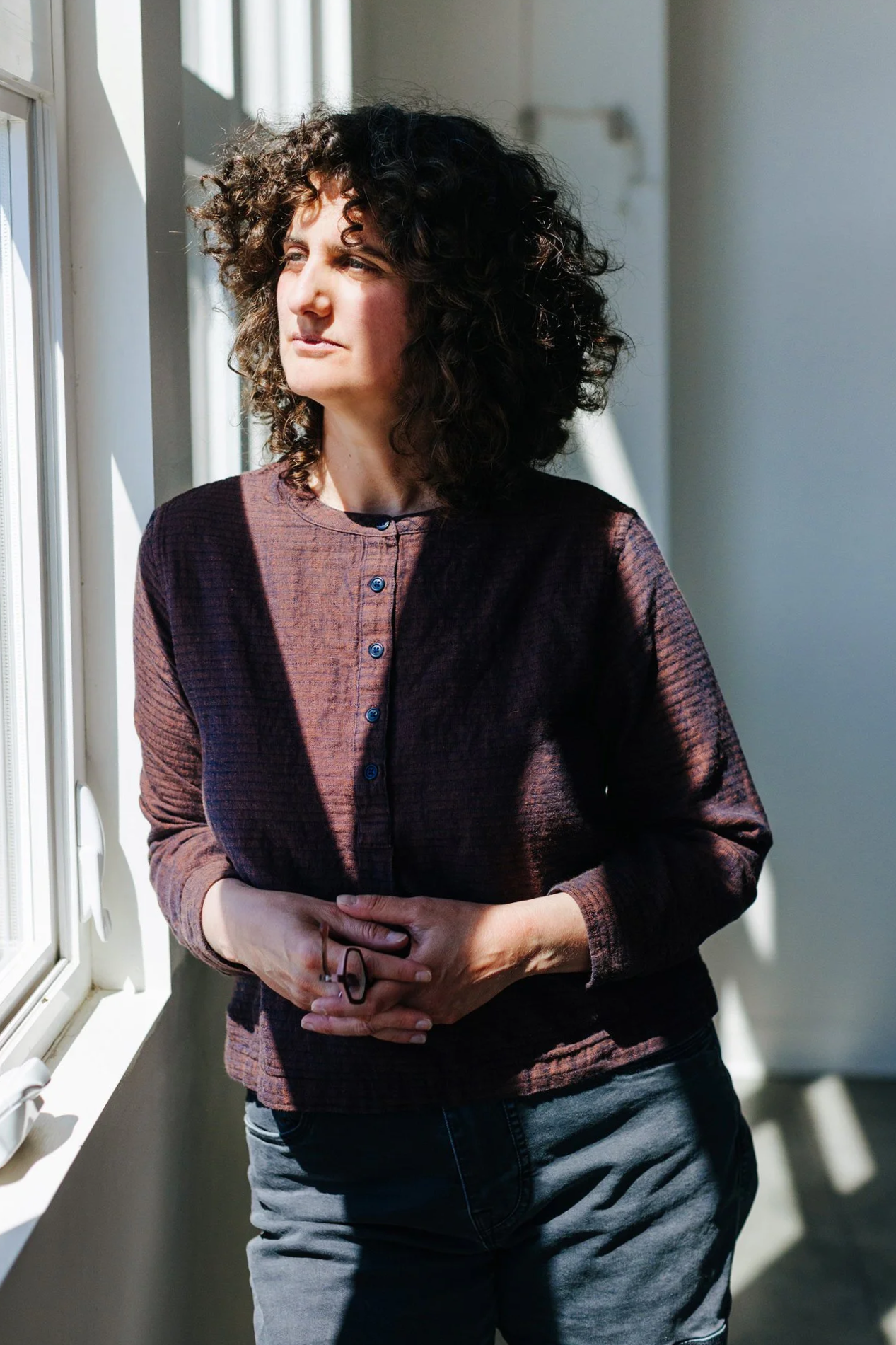 A woman with curly dark hair looking out a window with sunlight streaming in.