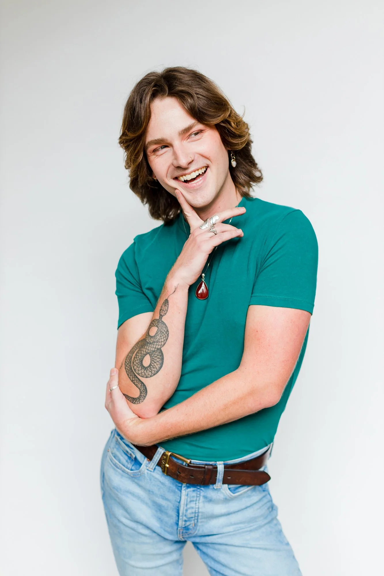 A young man with shoulder-length brown hair, wearing a teal t-shirt, light blue jeans, and a brown belt, smiling and posing with his hand on his chin. He has a tattoo of a snake on his left forearm, a silver ring on his right hand, and is wearing ear