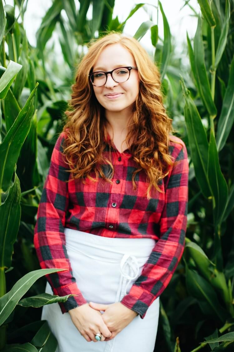A woman with red hair, glasses, wearing a red and black checkered shirt, standing among tall green cornstalks in a greenhouse or farm setting.