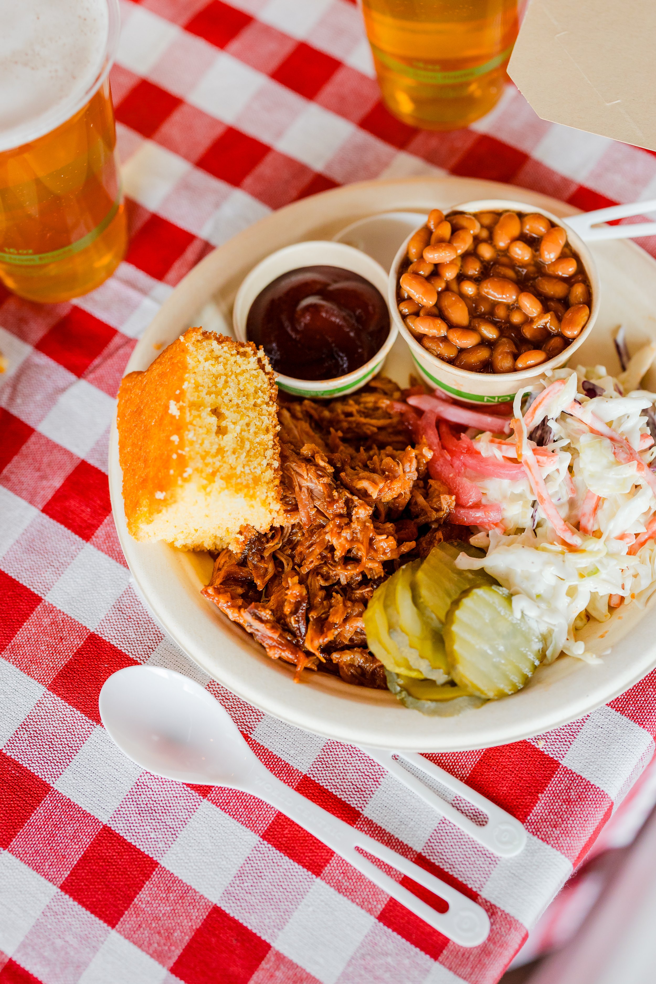 A plate of barbecue with coleslaw, pickles, baked beans, barbecue sauce, and cornbread, on a red and white checkered tablecloth with two glasses of beer.