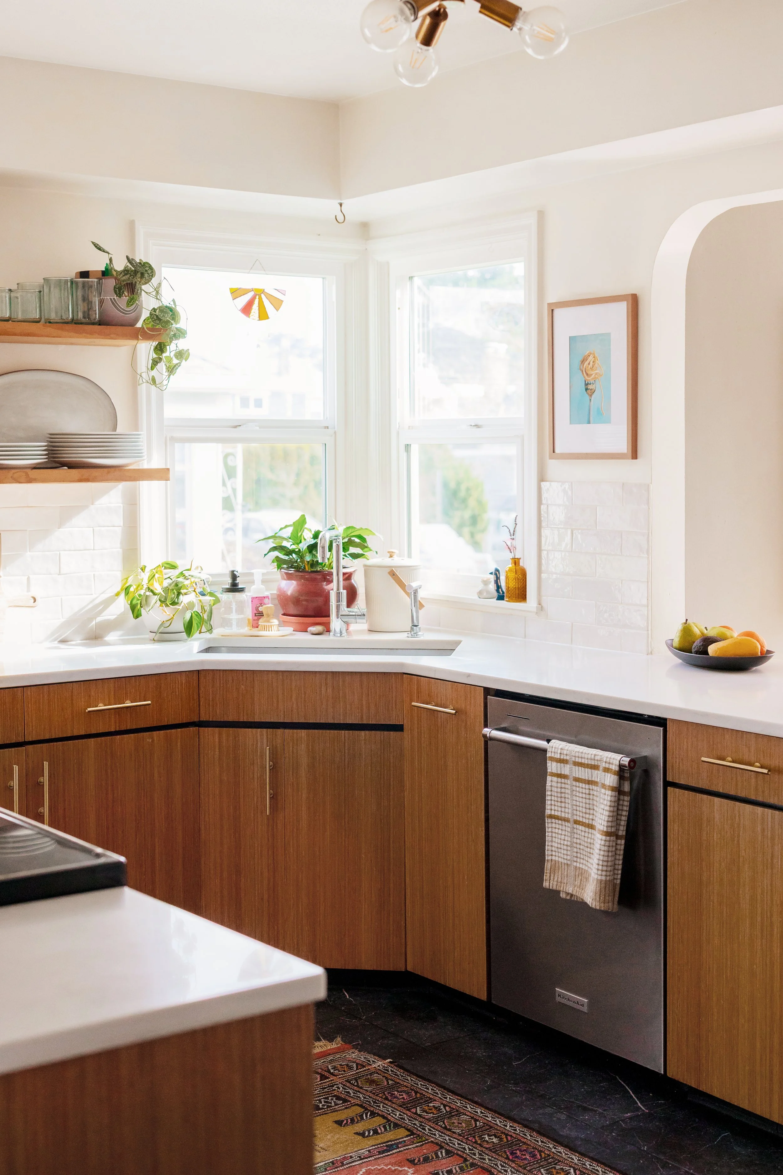Bright kitchen with white walls, large window, wooden cabinets, and a white countertop. Contains potted plants, bottles, and a framed artwork on the wall.