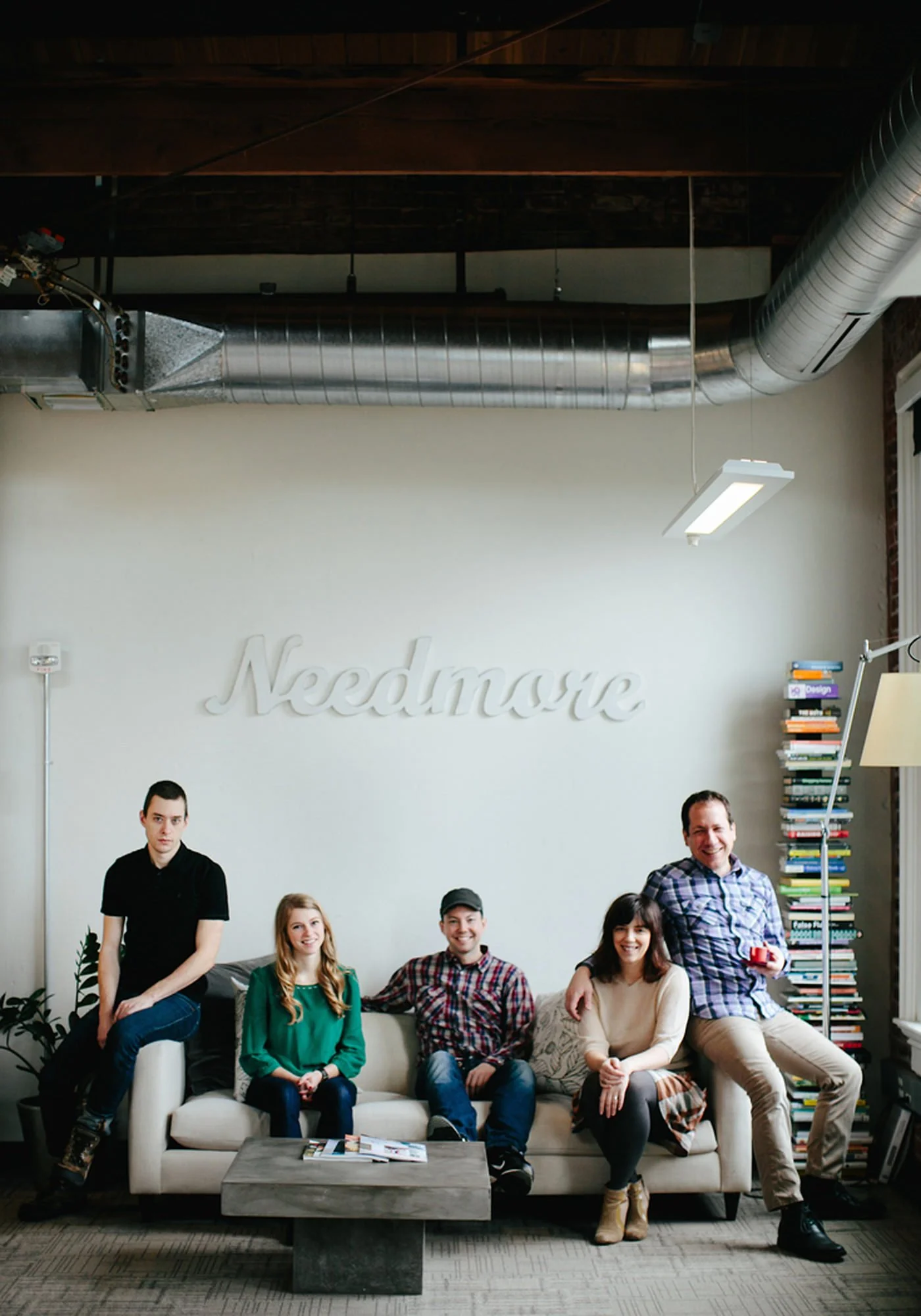 A group of five people sitting on a beige sofa in a modern office, with a white wall behind them displaying the word 'Nedmore'. The office has a bookshelf filled with multicolored books, a tall plant, and large windows, with an industrial ceiling fea