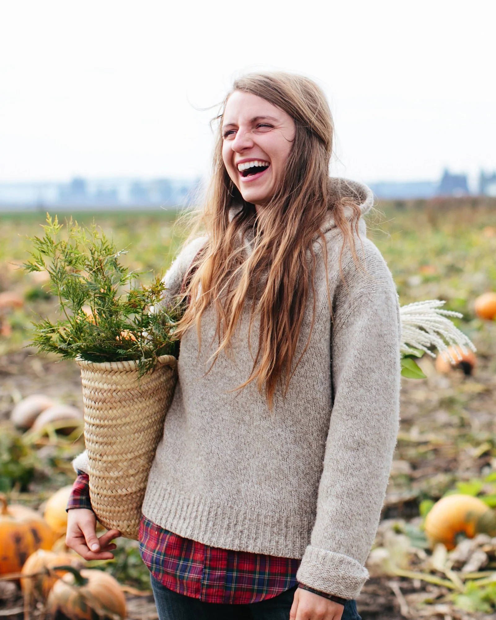 Young woman smiling in a pumpkin patch holding a woven basket with a plant inside, wearing a beige sweater and plaid shirt.