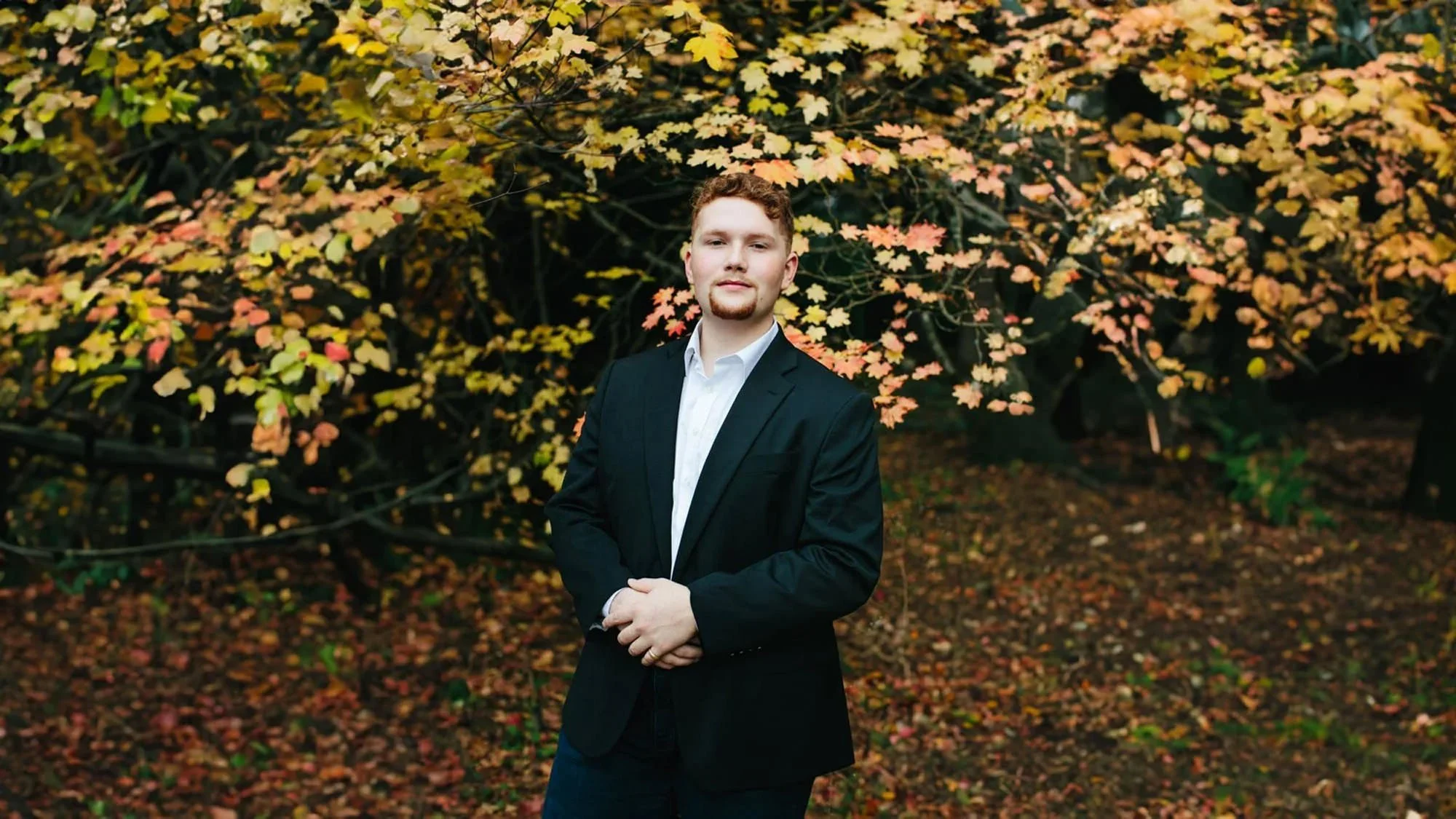 A young man with curly red hair and a beard stands outdoors in front of autumn foliage, wearing a black suit and white shirt, with his hands crossed in front of him.