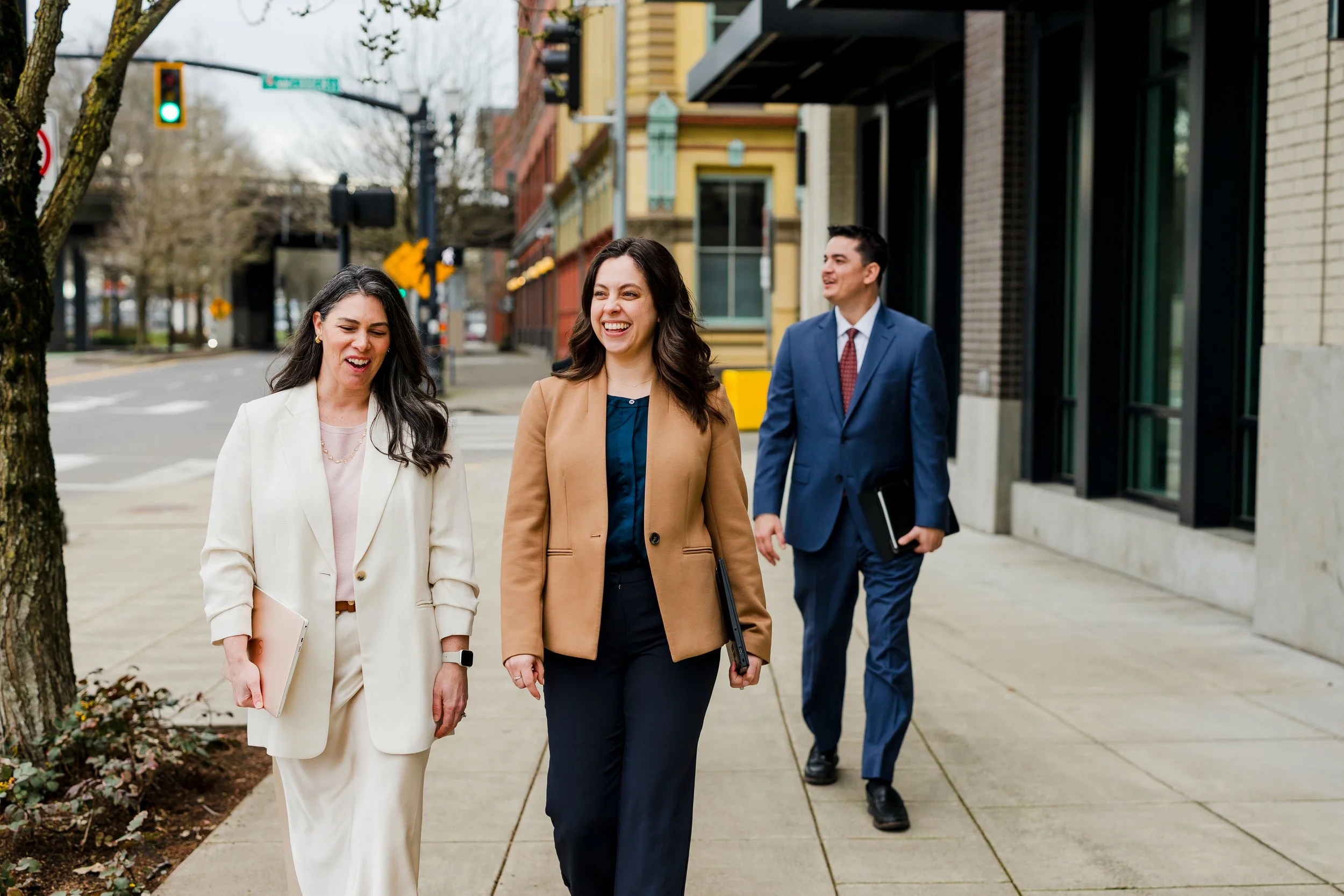 Three professionally dressed people walking on the sidewalk outdoors in a city, smiling and talking.