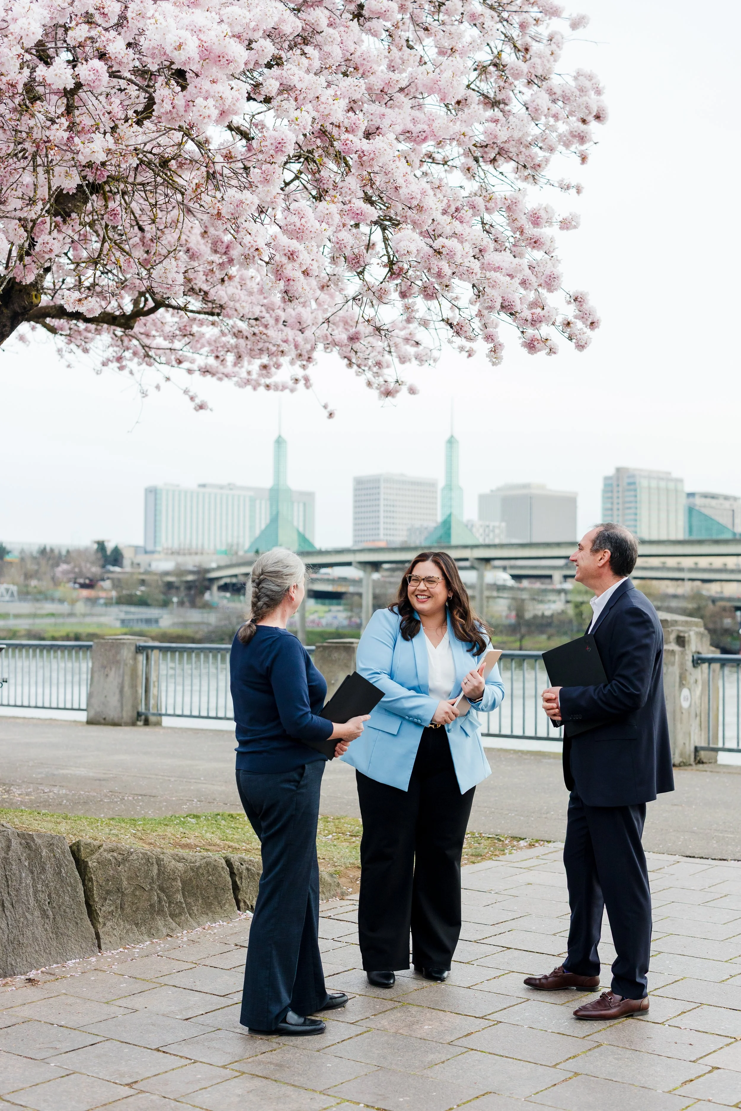 Three business professionals holding notebooks, standing and talking outdoors near a city river under a blooming cherry blossom tree.