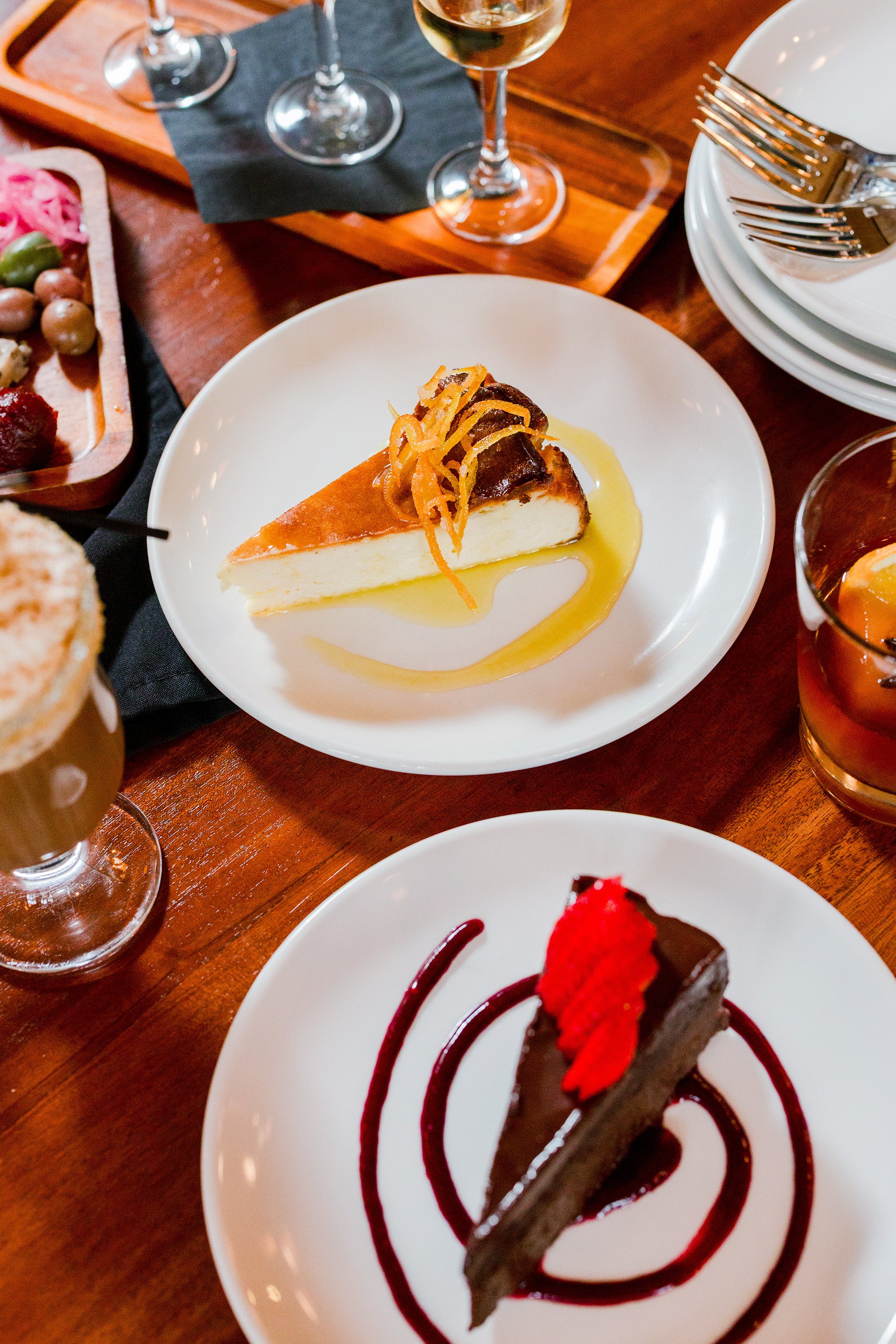 A table with a slice of cheesecake garnished with caramelized orange peels and sauce, and a slice of chocolate cake topped with a red berry, surrounded by drinks including wine, cocktails, and iced beverages.
