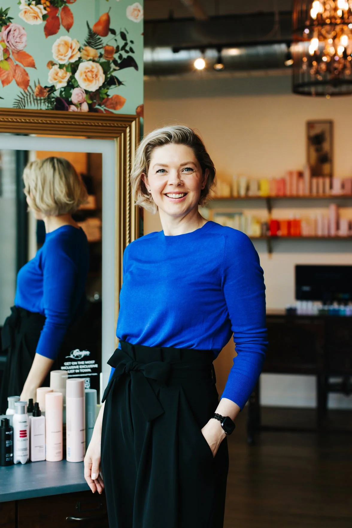 A smiling woman with short blonde hair, wearing a bright blue shirt and black high-waisted pants, stands in a well-lit room with a mirror and shelves of products in the background.