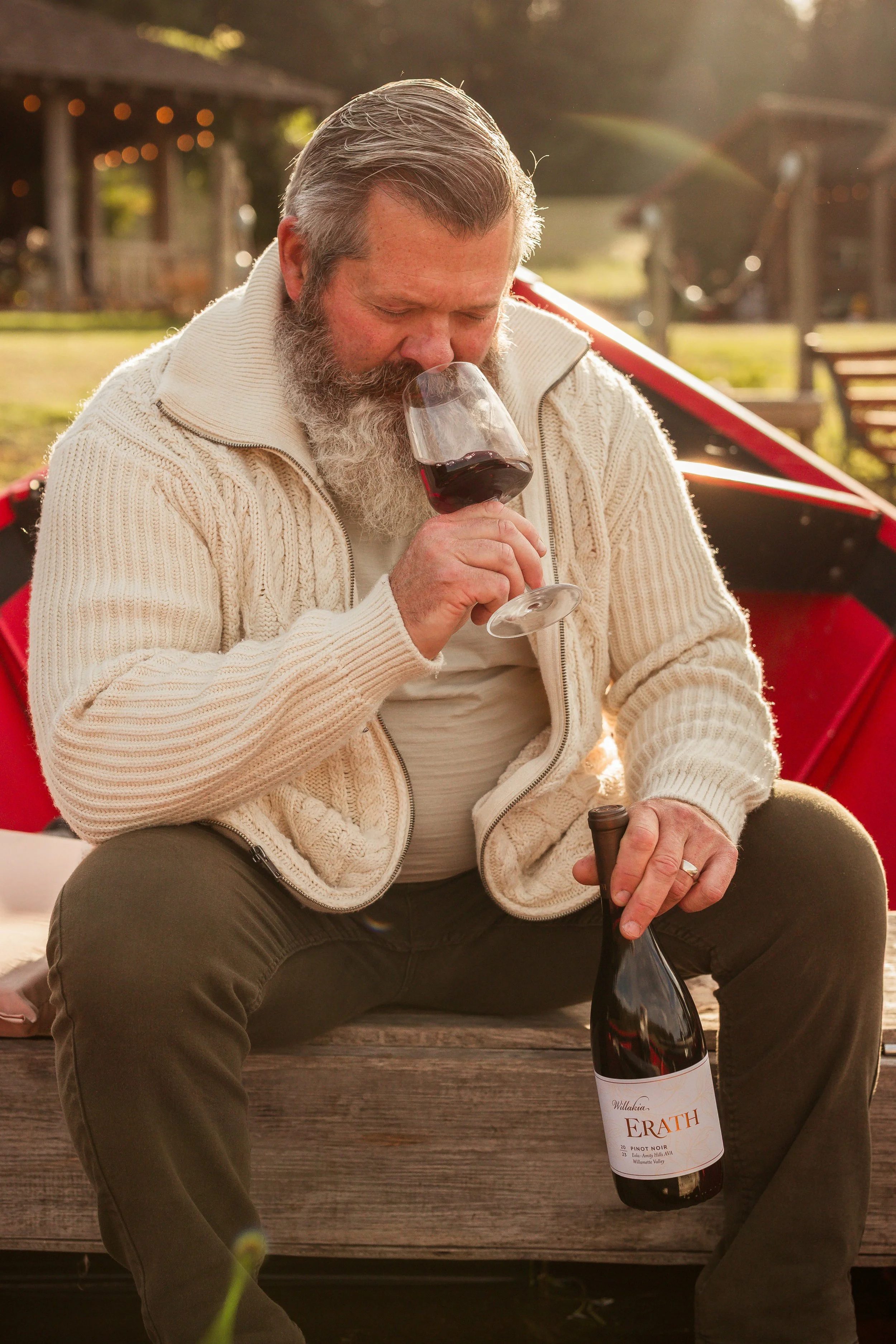 A man with a long gray beard, wearing a cream-colored knitted sweater, sitting on a wooden bench outdoors, sipping red wine from a glass, with a bottle labeled "ERATH" in his hand, during late afternoon sunlight.