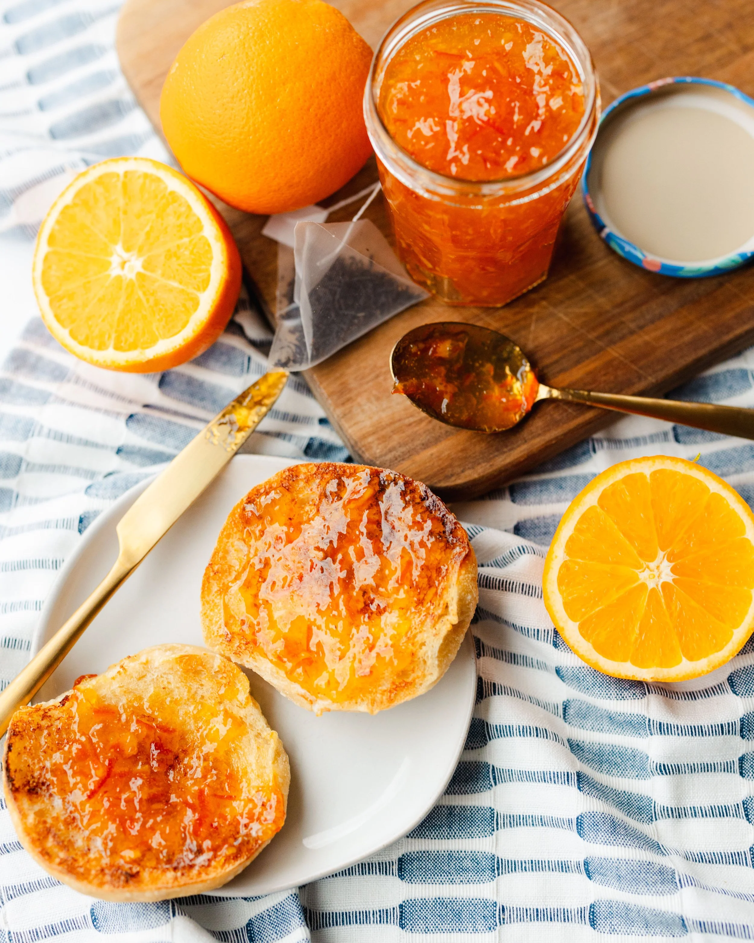 Half an orange, a jar of orange marmalade, a cup of milk, a spoon of marmalade, and toasted bread with marmalade on a white plate, all on a table with a striped blue and white cloth.