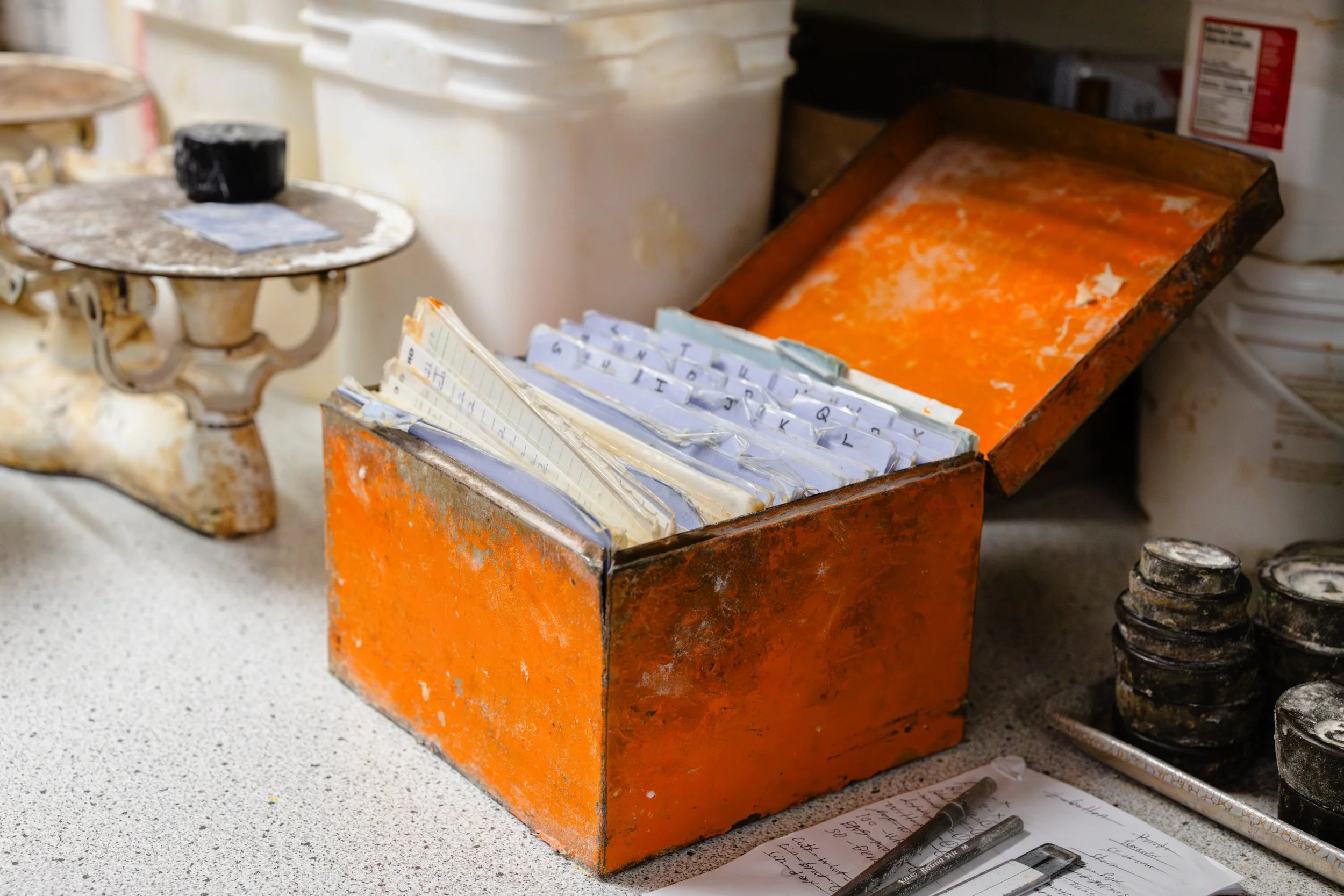 A cluttered workspace with an orange metal box containing a filing system, a small antique balance scale on the left, and various containers and papers scattered on a speckled countertop.