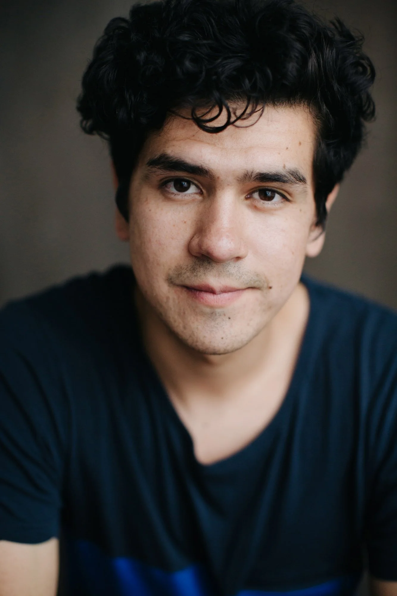 Close-up of a young man with dark, curly hair and light stubble, wearing a black t-shirt, looking at the camera with a slight smile.
