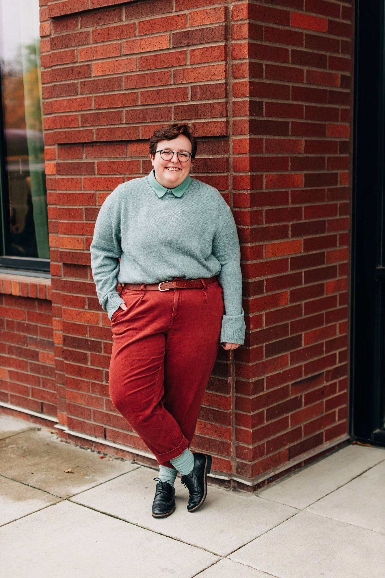 A person leaning against a brick wall smiling and wearing glasses, a light green sweater, a green collared shirt, red pants, light green socks, and black shoes.