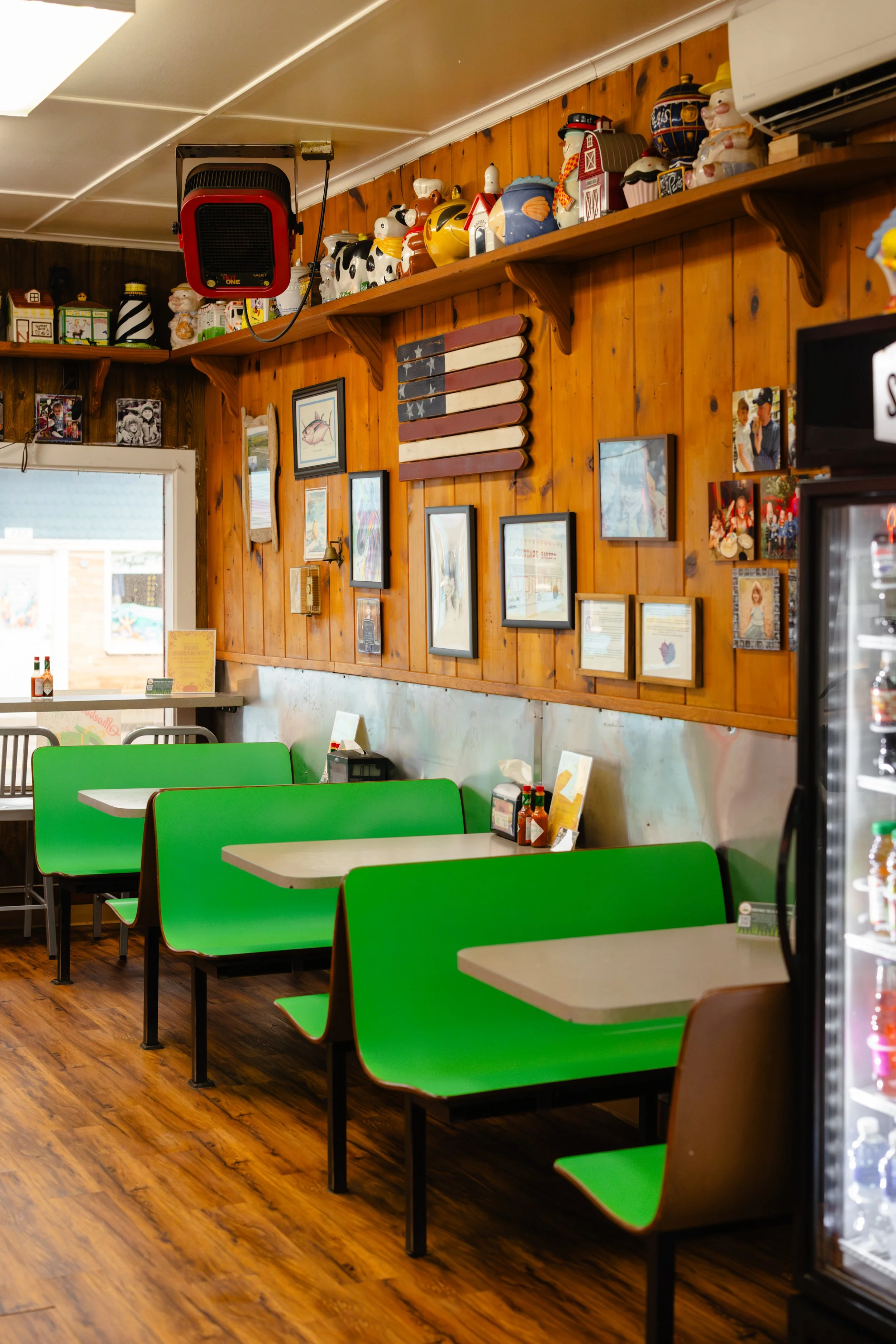 The interior of a casual restaurant with wooden walls and bright green booth seating. The walls are decorated with framed pictures, a small American flag made of wooden planks, and various knickknacks, including ceramic and figurine items on a shelf 