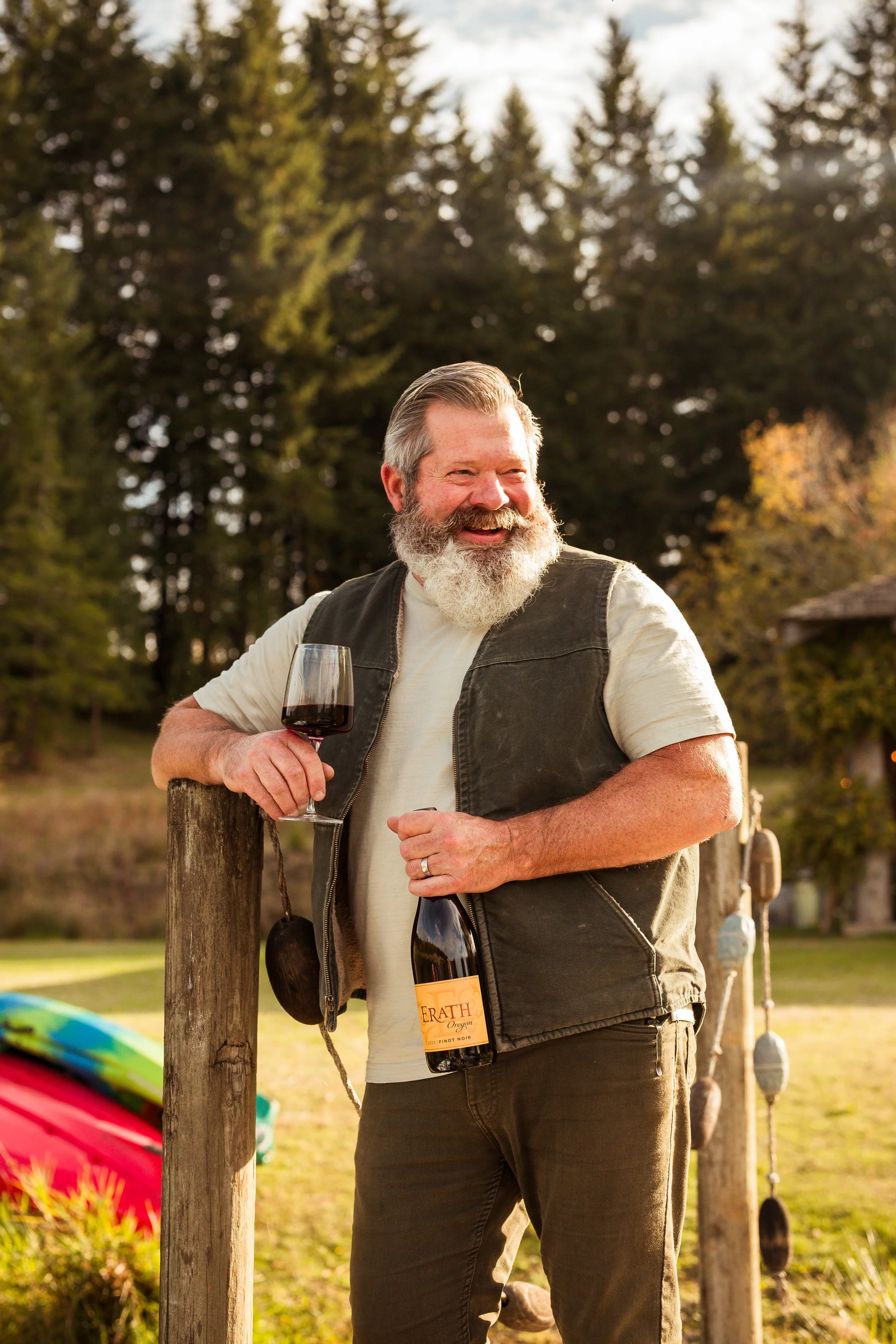 An older man with a gray beard and hair, wearing a white t-shirt and a black vest, is smiling and holding a glass of red wine and a bottle in an outdoor setting with trees and a fence.