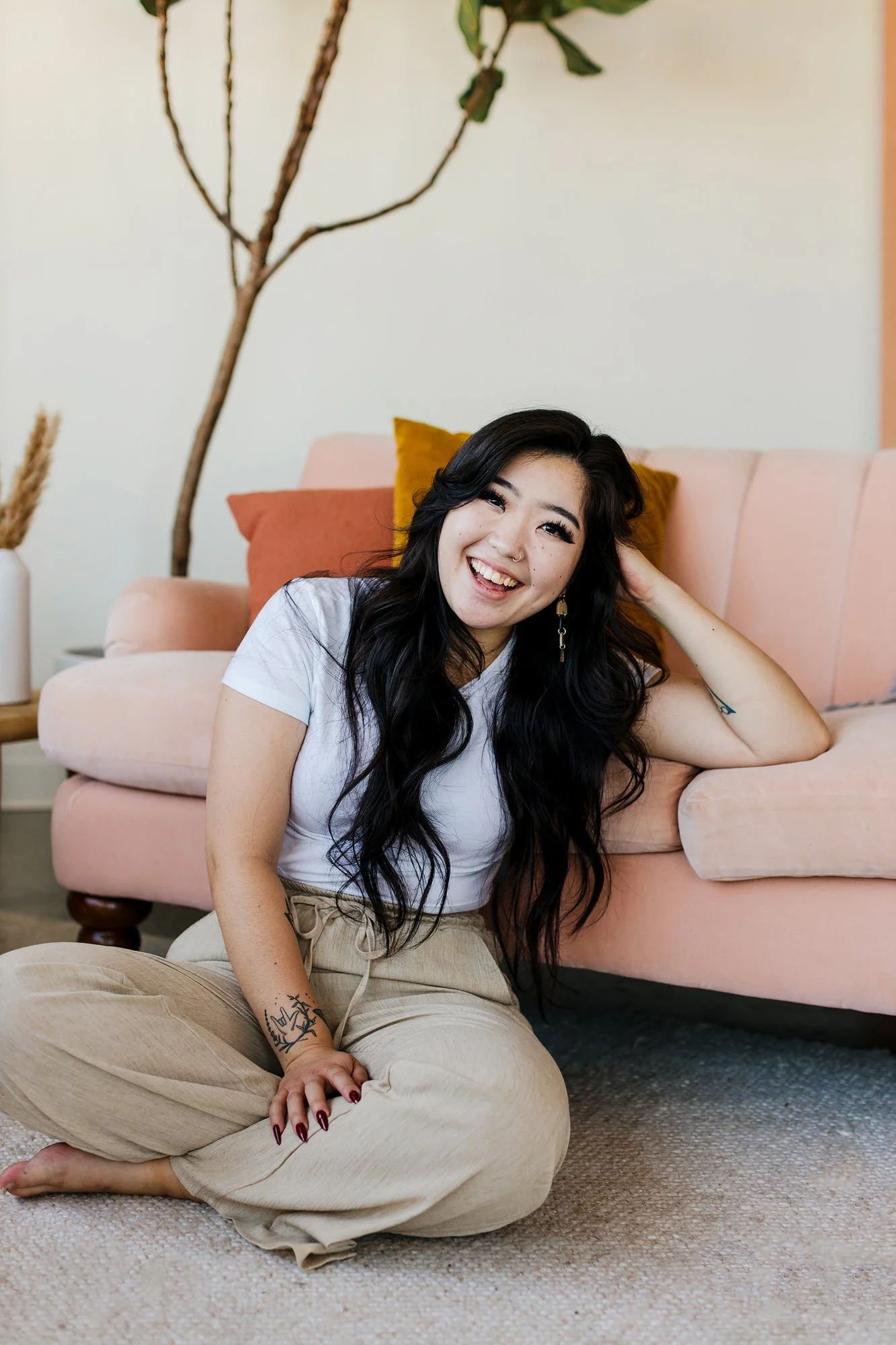 A woman with long black wavy hair sitting on the floor next to a pink sofa, smiling and leaning her head on her hand. She is wearing a white t-shirt and beige pants, with a tattoo on her arm. There is a mustard yellow pillow and a rust-colored pillow