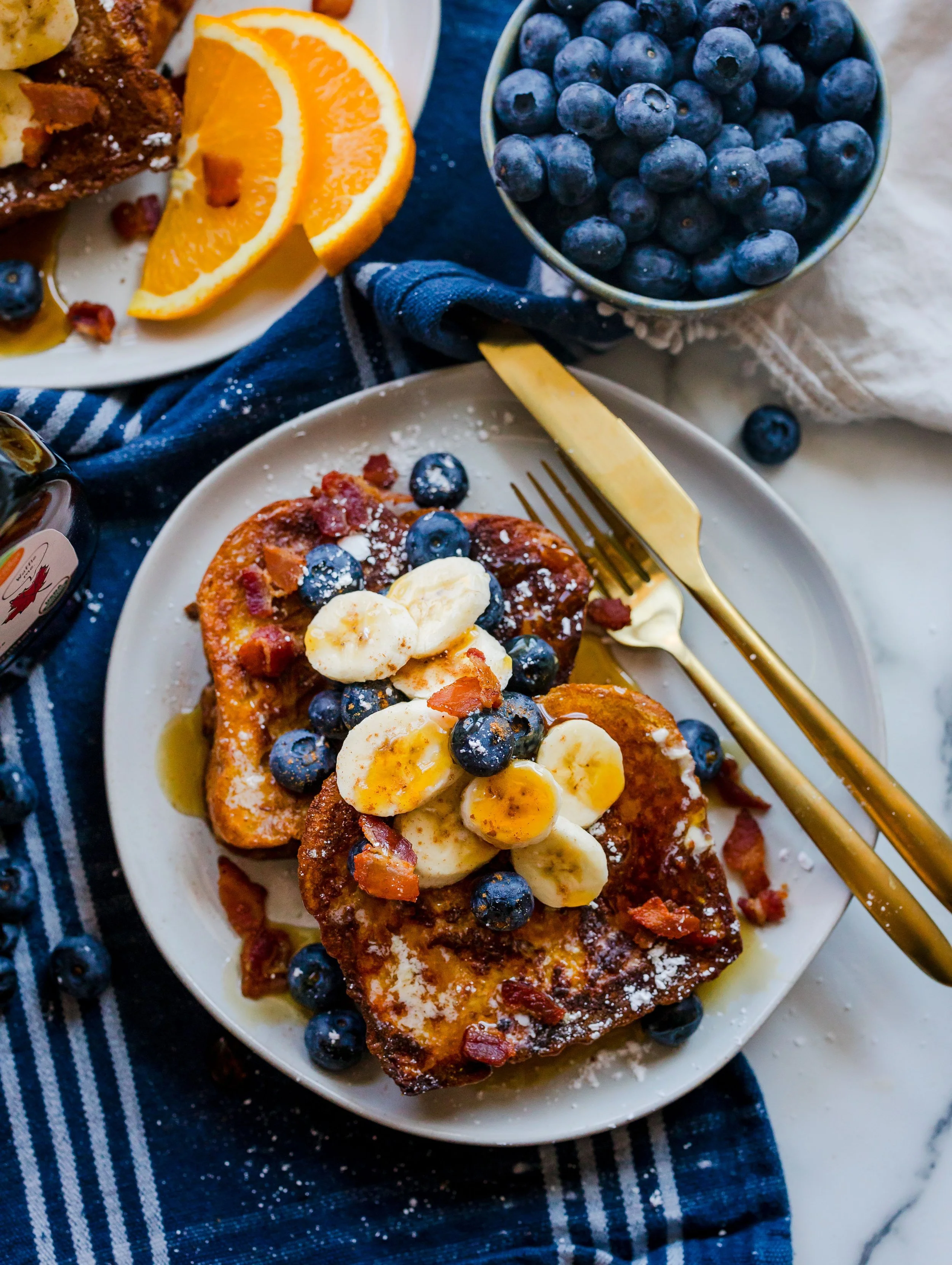 French toast topped with sliced bananas, blueberries, bacon, powdered sugar, and syrup on a white plate.