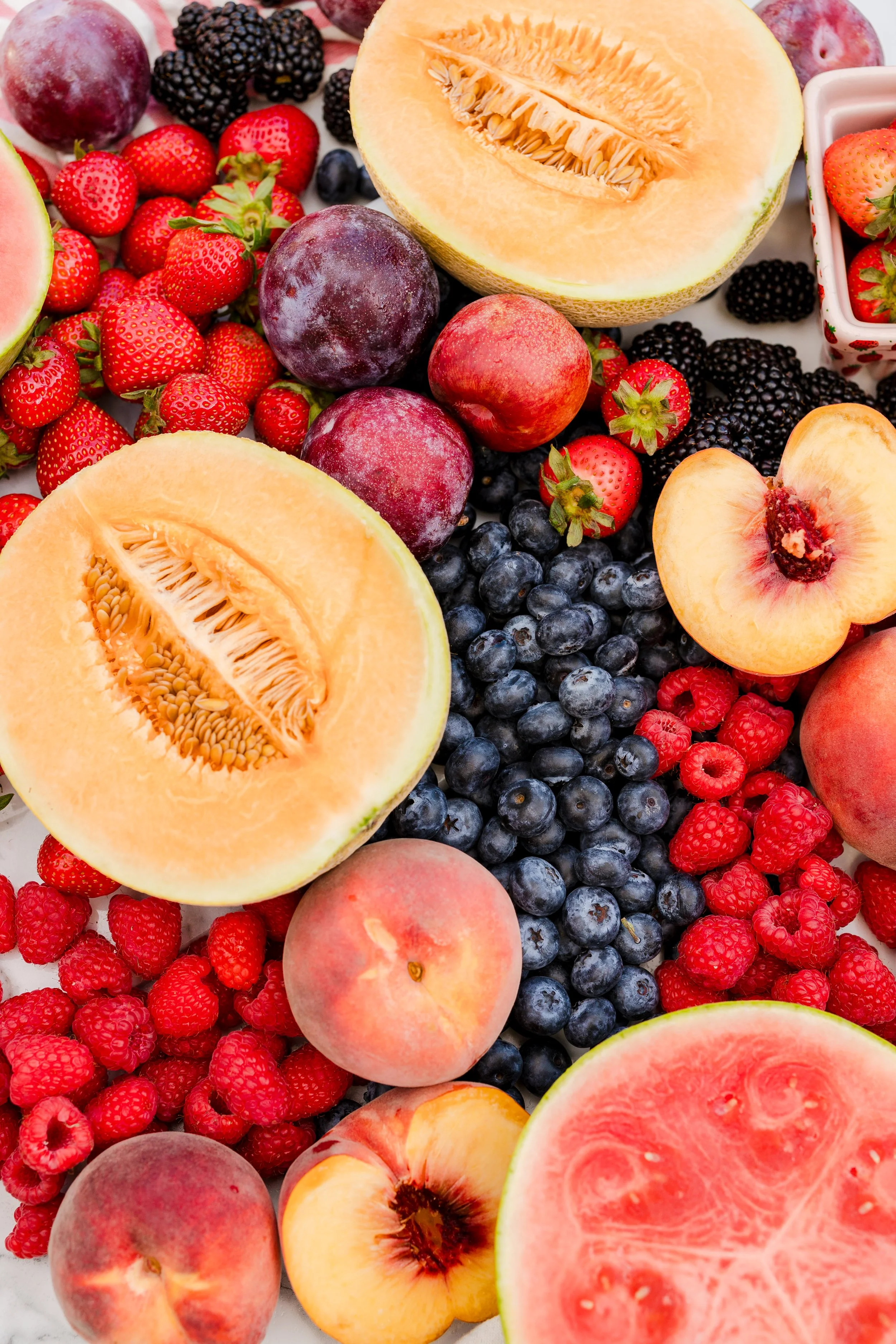 A variety of fresh fruits including halved cantaloupe, watermelon slices, peaches, strawberries, blackberries, blueberries, and raspberries arranged on a surface.