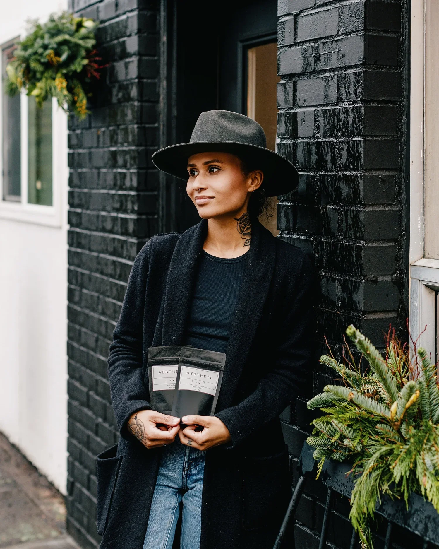 A woman leaning against a black brick wall, wearing a black coat, dark wide-brimmed hat, and holding two black packages labeled 'AESTHETE'. She has a tattoo on her neck and one hand has a tattoo, and there is a window and a decorated bush nearby.
