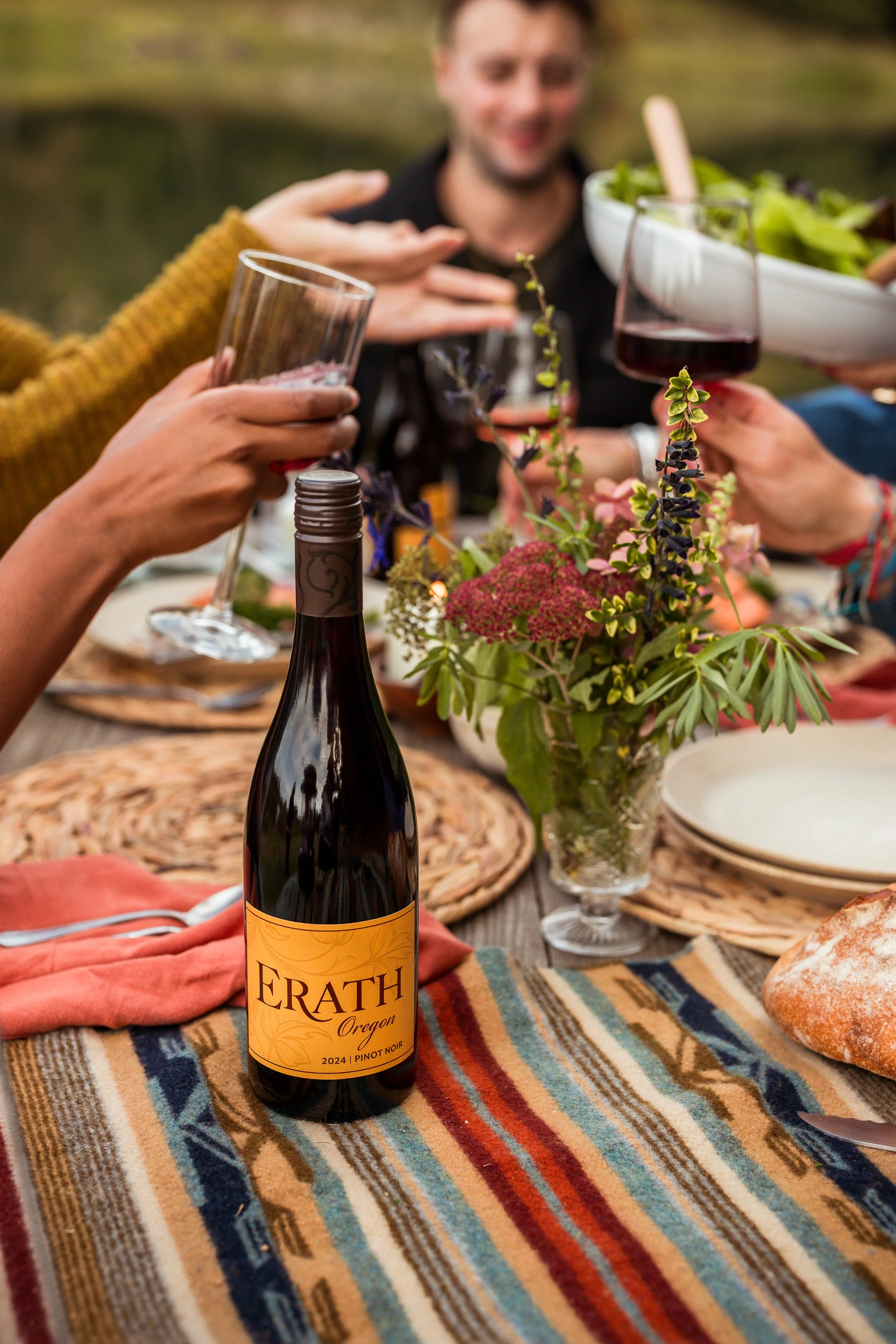 A group of people celebrating outdoors around a table with a bottle of Erath Oregon Pinot Noir wine, glasses of red wine, a bowl of salad, and a bouquet of flowers, with a smiling man in the background.