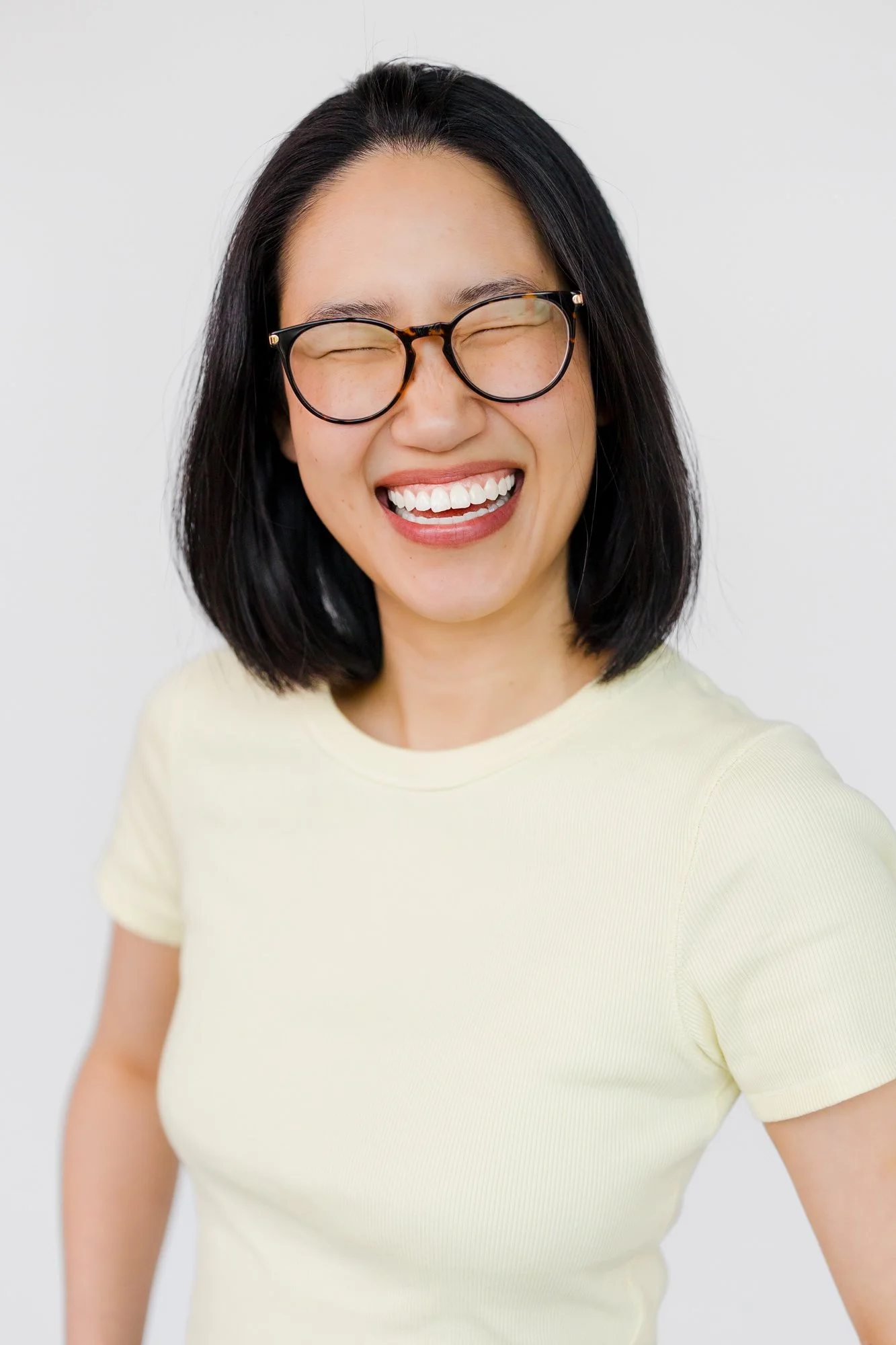 A woman with black hair and glasses is smiling and laughing, wearing a light-colored shirt, against a plain white background.