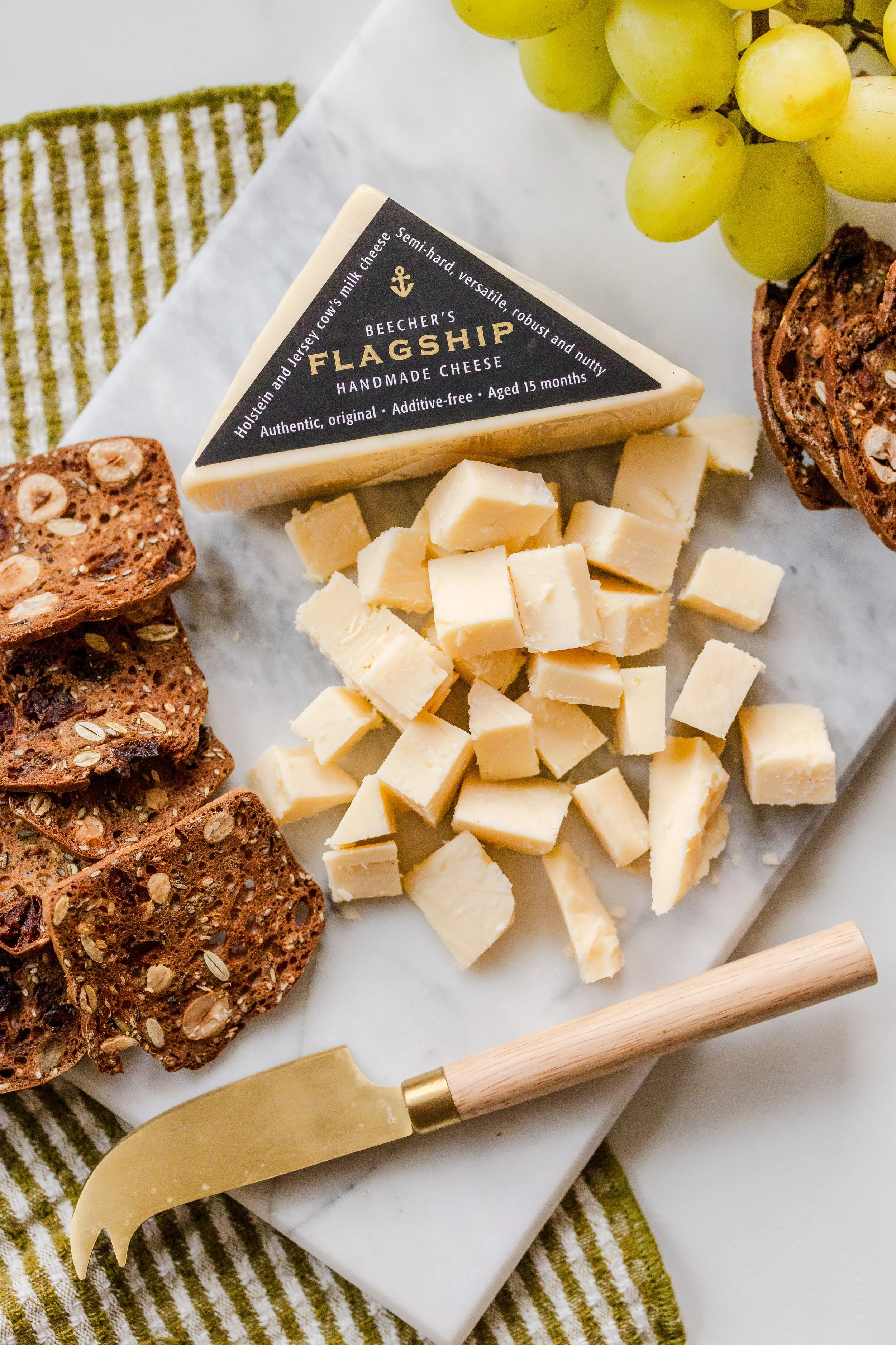 A wedge of Beechers Flagship semi-hard cheese, cubed pieces of cheese, sliced multigrain crackers with oats and seeds, a bunch of green grapes, a cheese knife with a wooden handle, all on a white marble cutting board with a green striped cloth undern