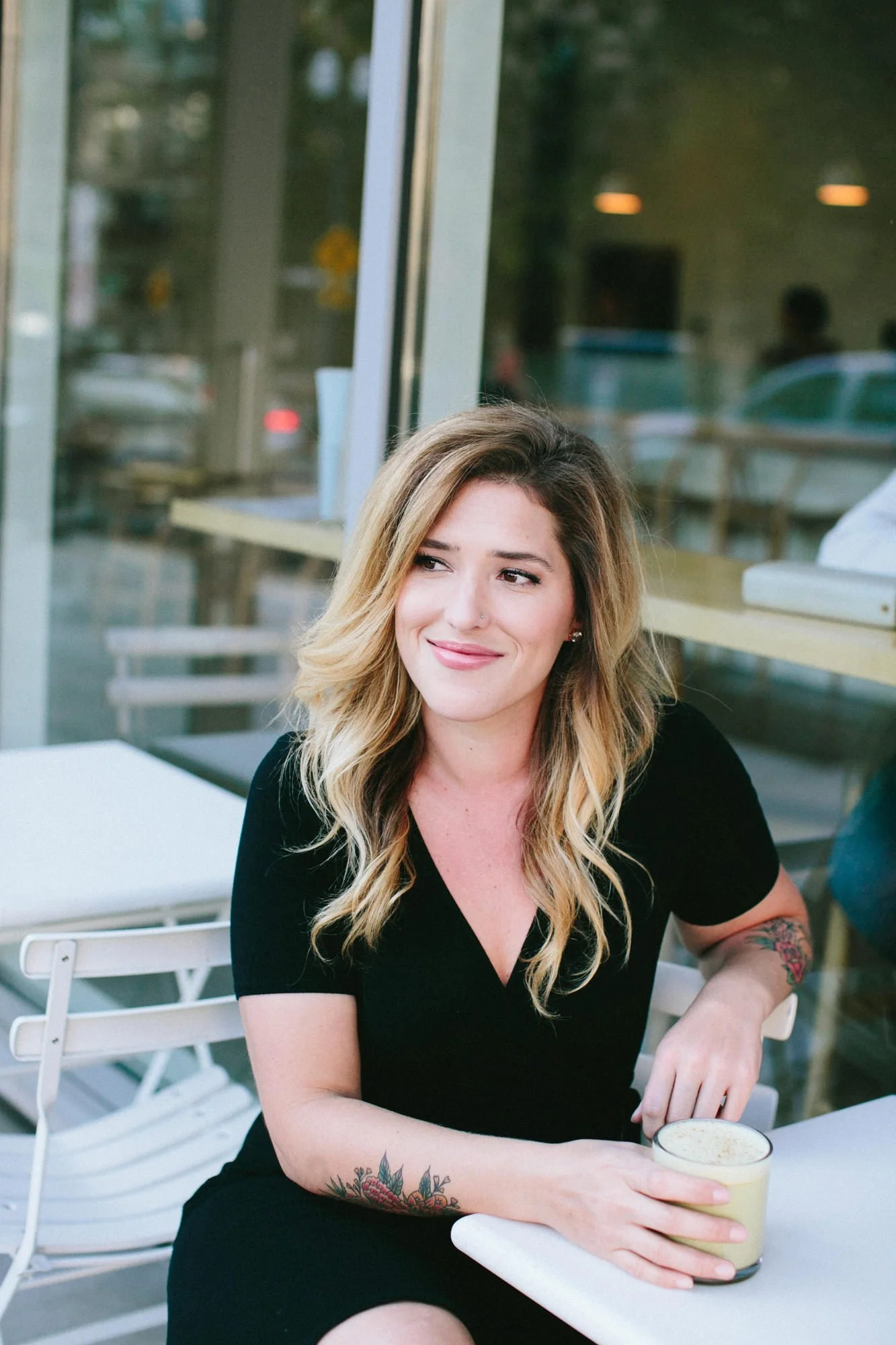 A woman with blonde, wavy hair and tattoos on her arm, wearing a black shirt, sitting at an outdoor cafe table with a drink in her hand, smiling.