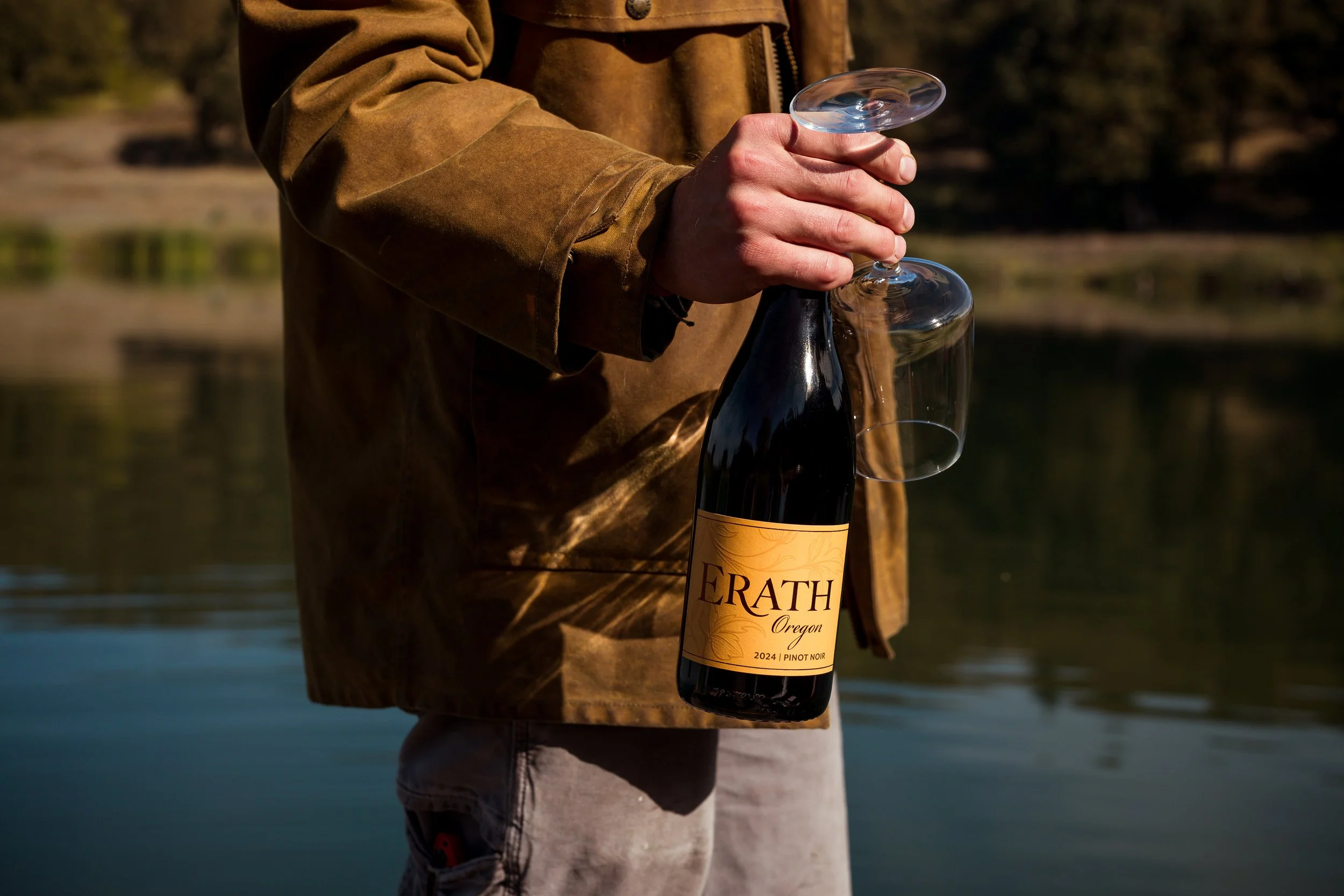 Person holding a bottle of Erath Oregon Pinot Noir 2024 wine and two empty wine glasses outdoors.