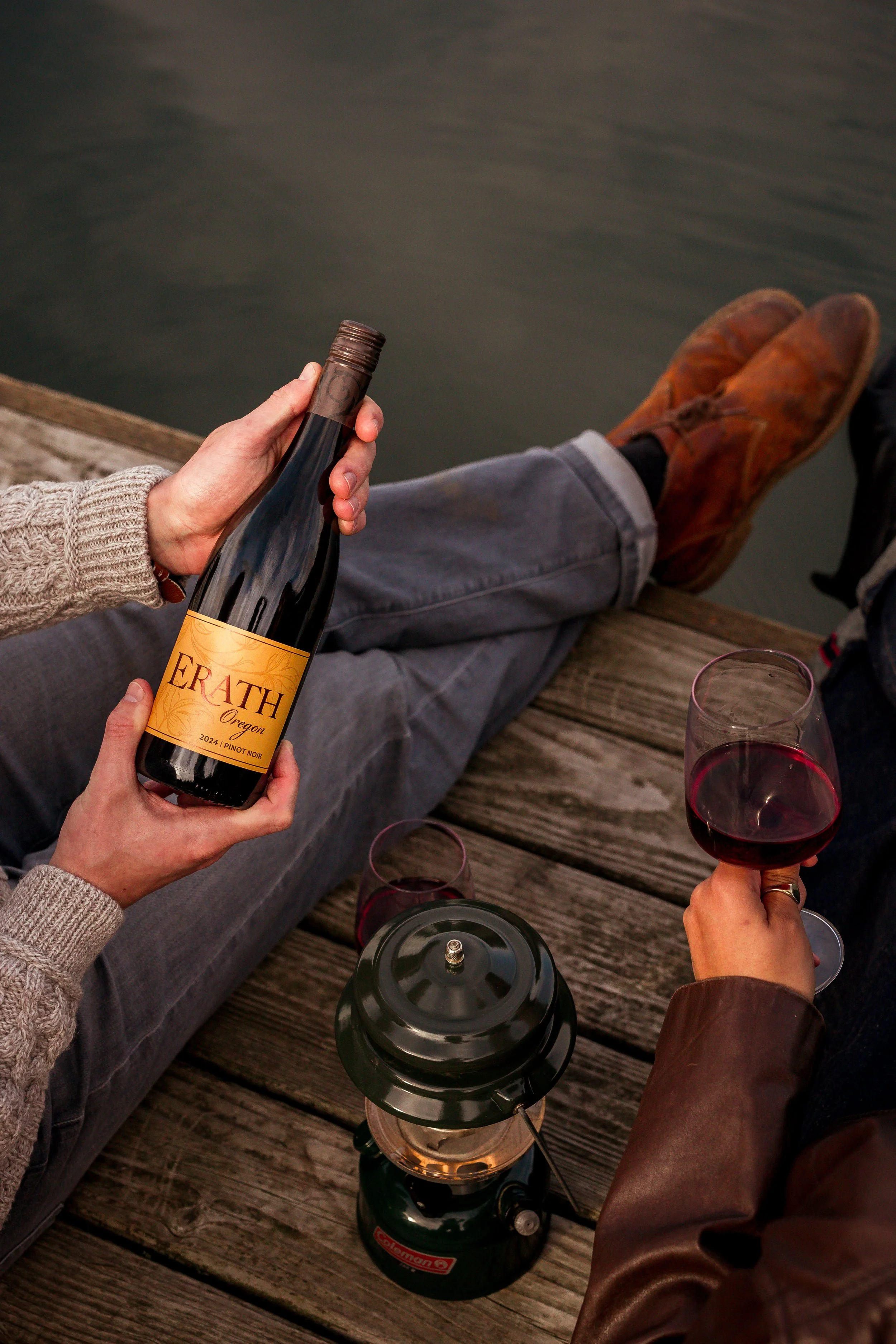 Two people sitting on a wooden dock by the water, enjoying red wine and a bottle of Erath Oregon Pinot Noir. One person holds the wine bottle, and the other holds a wine glass. A lantern is also on the dock.
