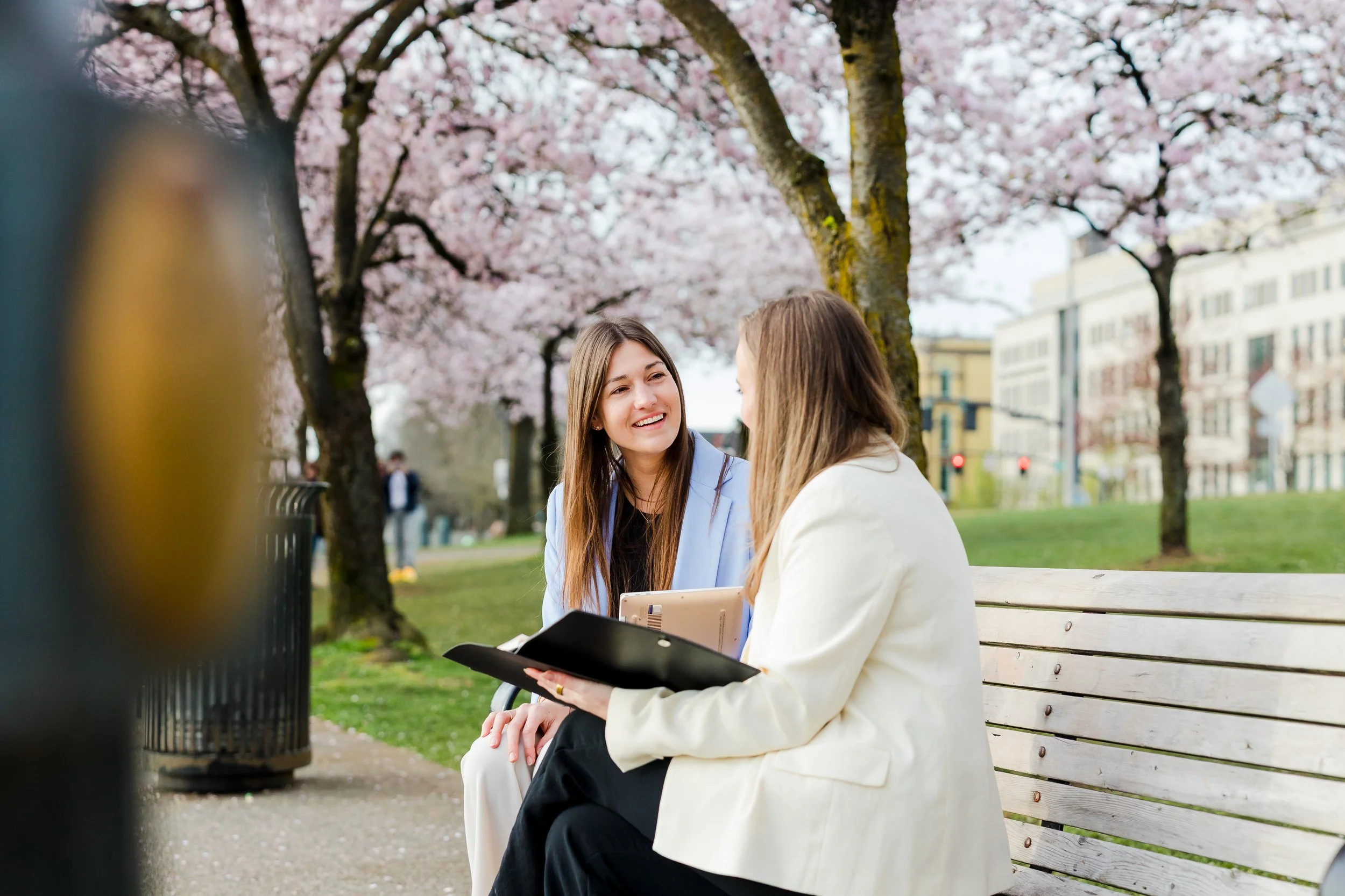 Two women sitting on a park bench under blooming cherry blossom trees, smiling and talking, with city buildings in the background.