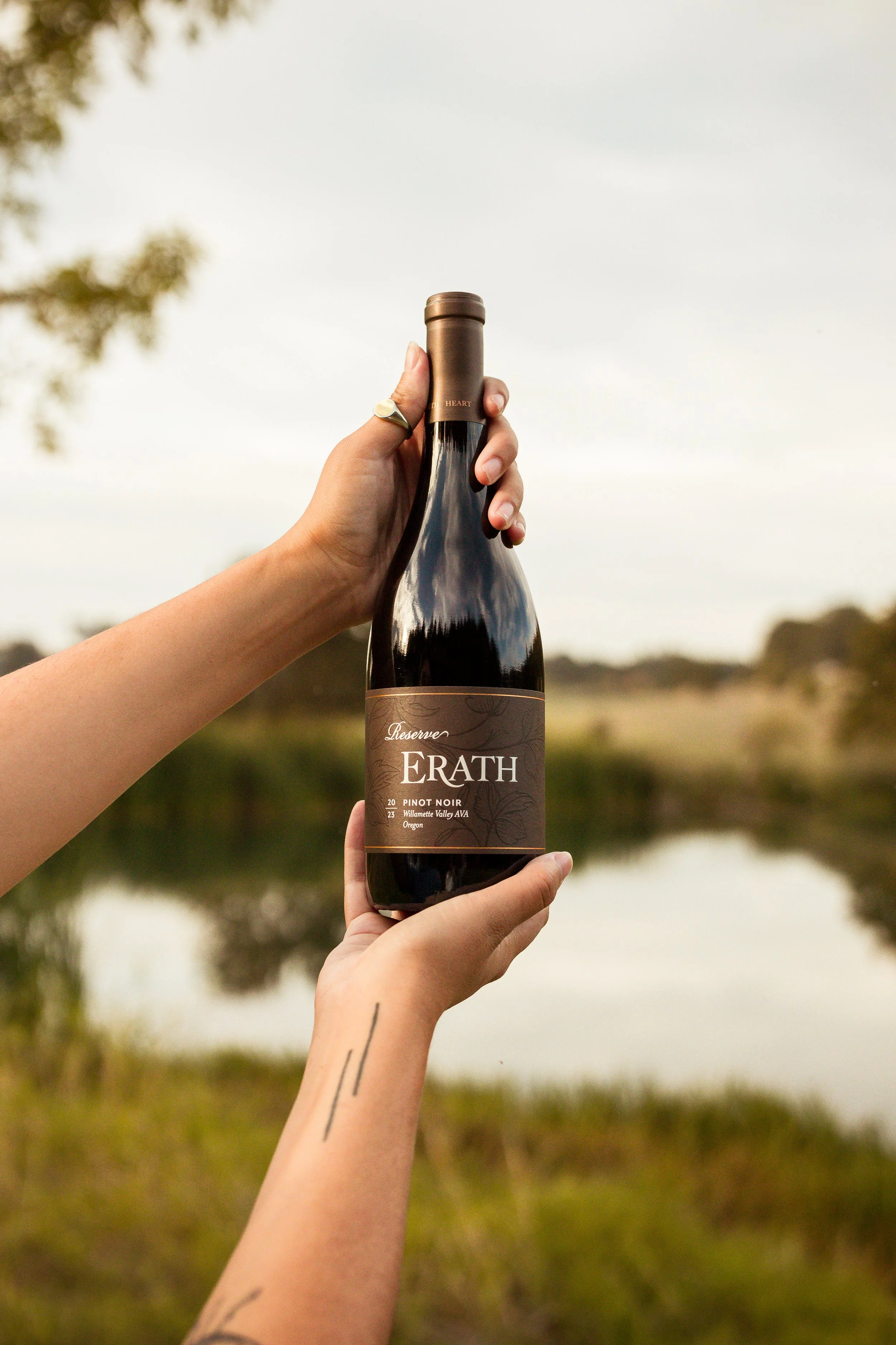 A person holding a bottle of Erath Pinot Noir wine outdoors near a body of water, with a scenic background of trees and cloudy sky.