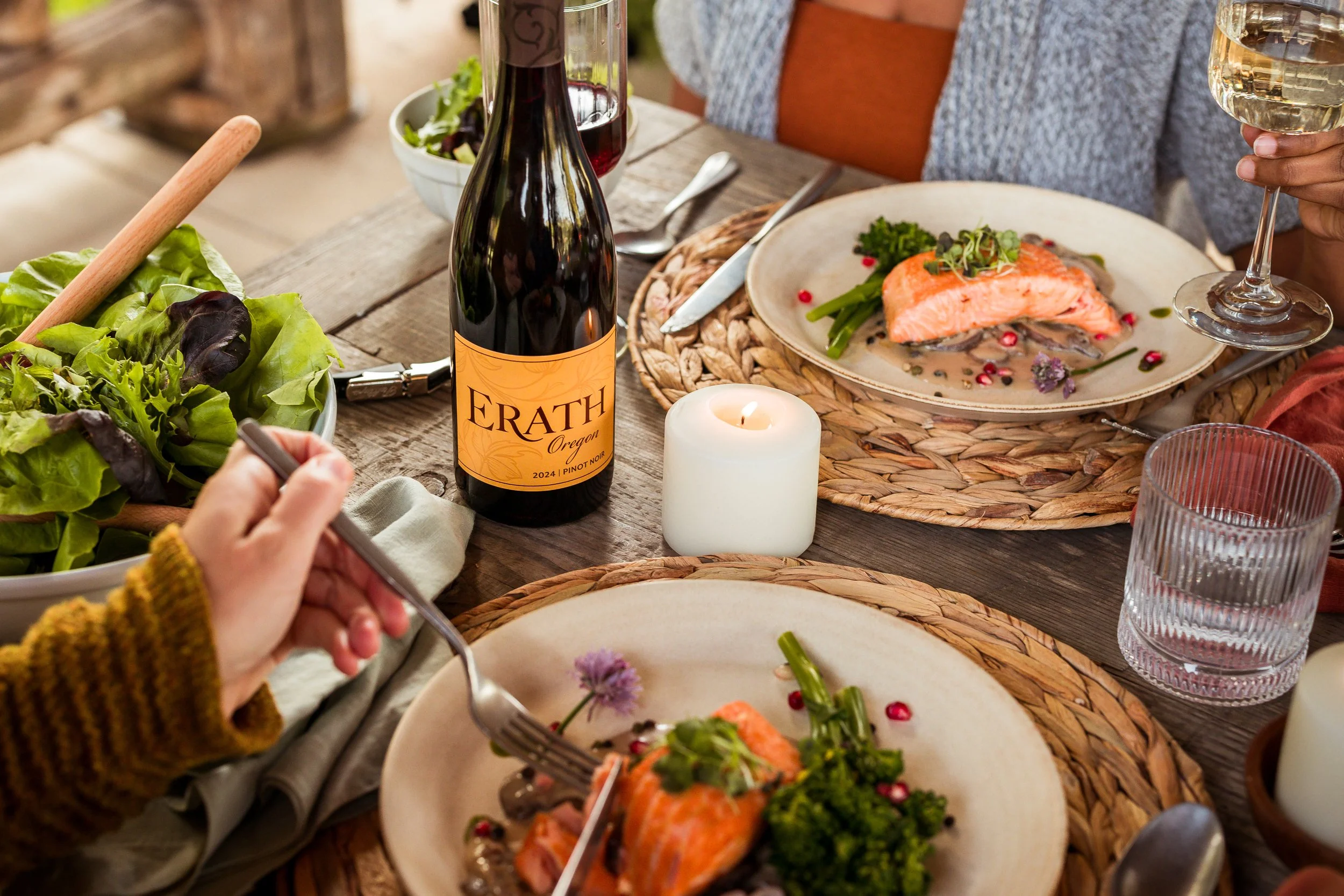 Family gathering around a rustic wooden table enjoying a meal with plates of salmon, green vegetables, wine, and a lit candle, with a bottle labeled 'Erath Oregon' on the table.